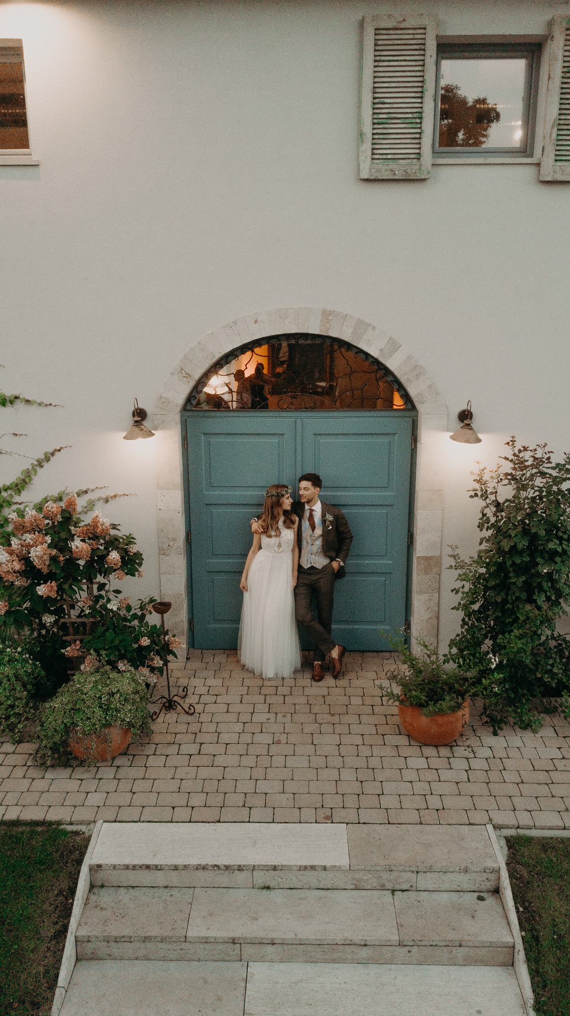 Para młoda w stylizowanych strojach pozuje na tle błękitnych drzwi z kamiennym łukiem i roślinami w donicach. Ujęcie z góry, ciepłe oświetlenie.