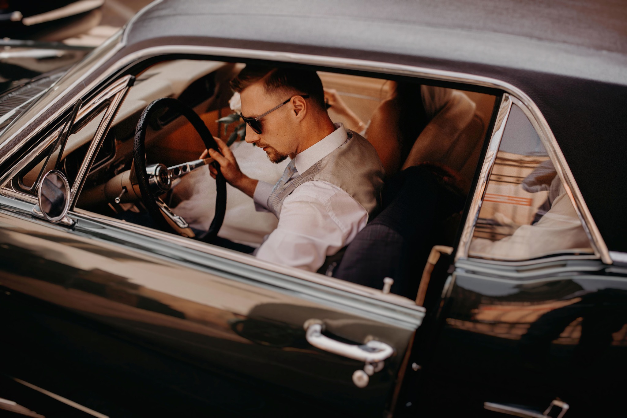 Elegancki mężczyzna w okularach przeciwsłonecznych siedzi za kierownicą klasycznego, ciemnozielonego samochodu. Ubrany w białą koszulę i kamizelkę.