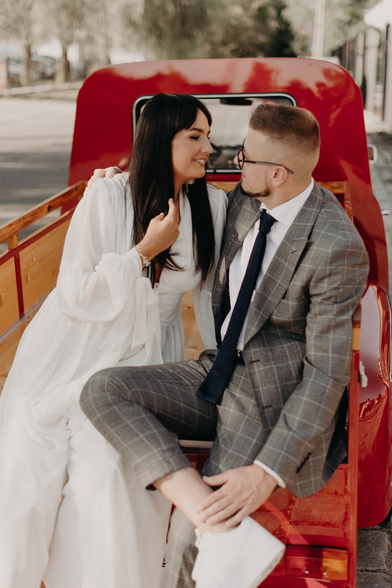 Uśmiechnięta para w stylizowanych strojach siedzi w czerwonym, zabytkowym samochodzie, spoglądając na siebie podczas sesji plenerowej.