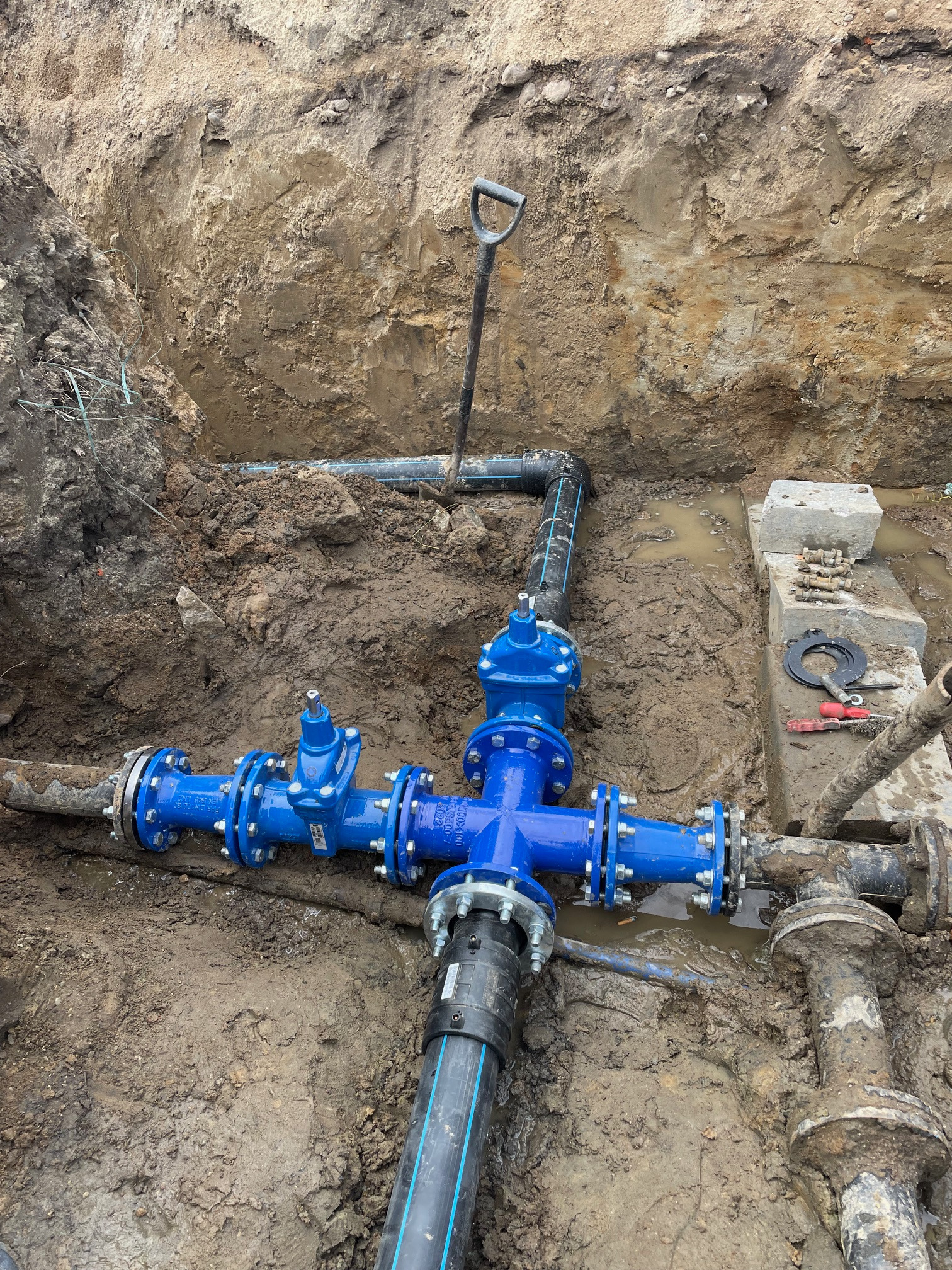 Niebieskie rury z zaworami w wykopie, widoczne narzędzia i fragment ściany wykopu. Etap prac instalacyjnych przyłącza wodno-kanalizacyjnego.