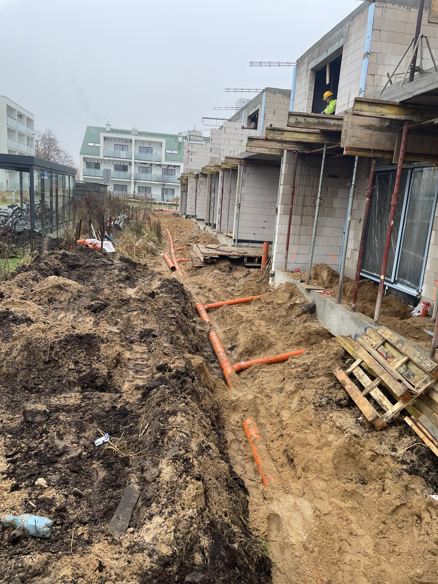 Wykop z ułożonymi pomarańczowymi rurami kanalizacyjnymi na tle budynków w trakcie budowy. Widoczny pracownik w żółtym kasku. Paleta i narzędzia obok wykopu.