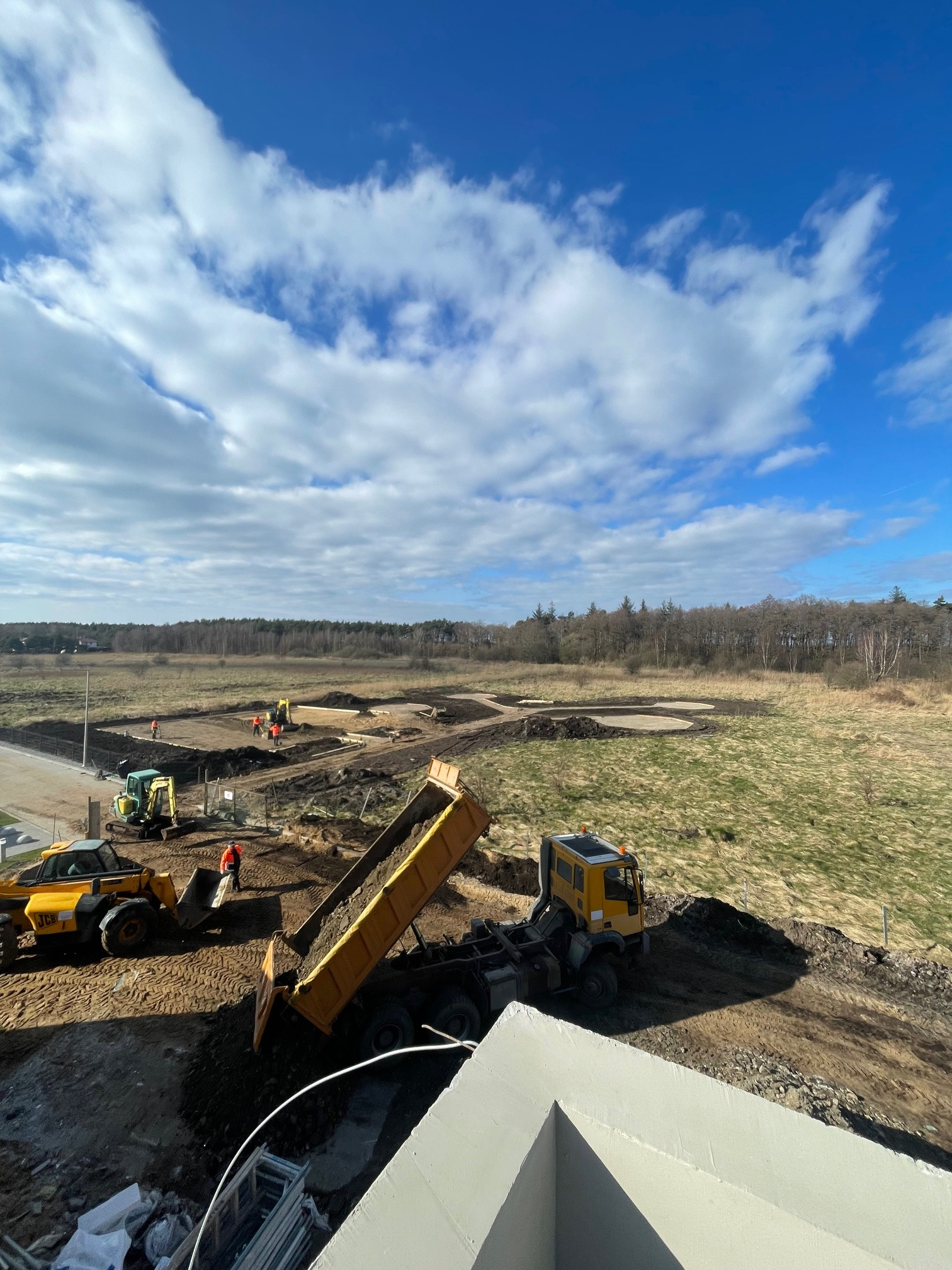 Żółty wywrotka wysypuje ziemię na plac budowy z pracującymi maszynami i robotnikami w oddali, błękitne niebo z chmurami w tle. Widok z góry.