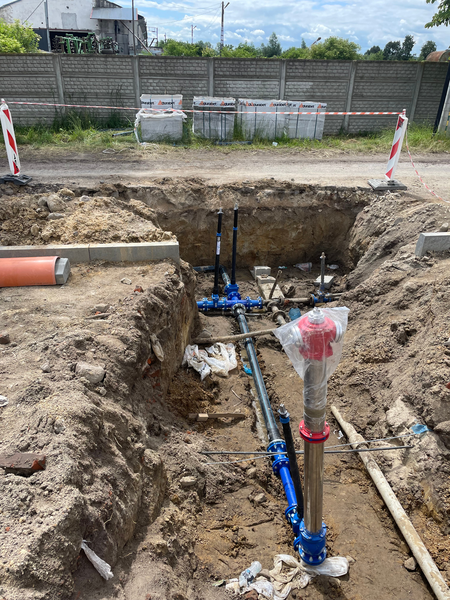 Wykop z widoczną, częściowo zamontowaną instalacją wodociągową z hydrantem w osłonie. W tle ogrodzenie z betonowych płyt i materiały budowlane.