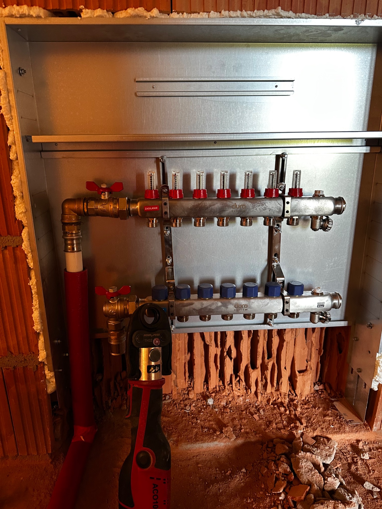 Rozdzielacz ogrzewania podłogowego 'duro' w metalowej szafce, widoczna praska hydrauliczna 'novopress' i fragment instalacji w surowej cegle.