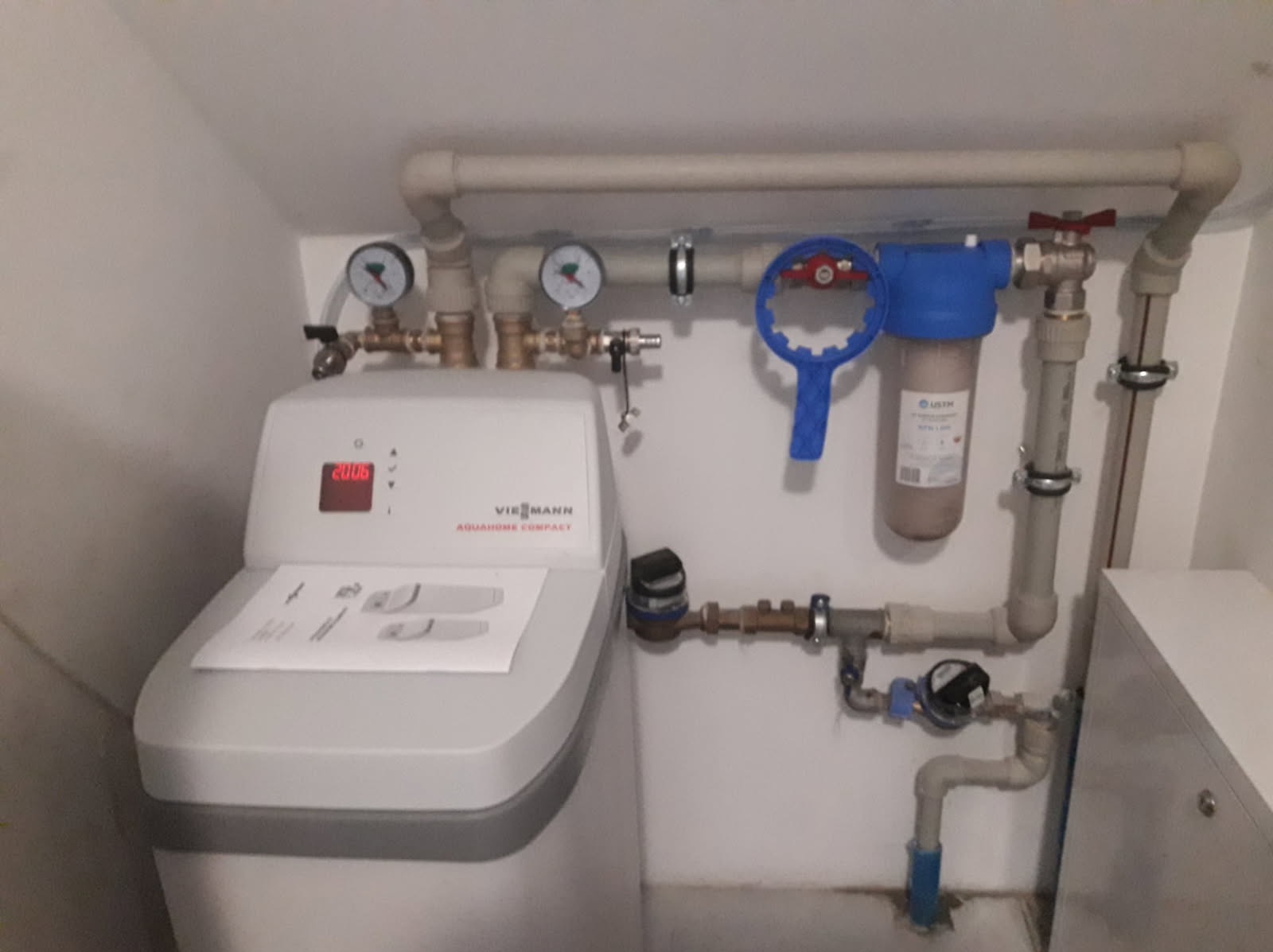 Zamontowany zmiękczacz wody Viessmann Aquahome Compact z manometrami, filtrem narurowym i wodomierzem w białym pomieszczeniu technicznym, z rurami PCV.