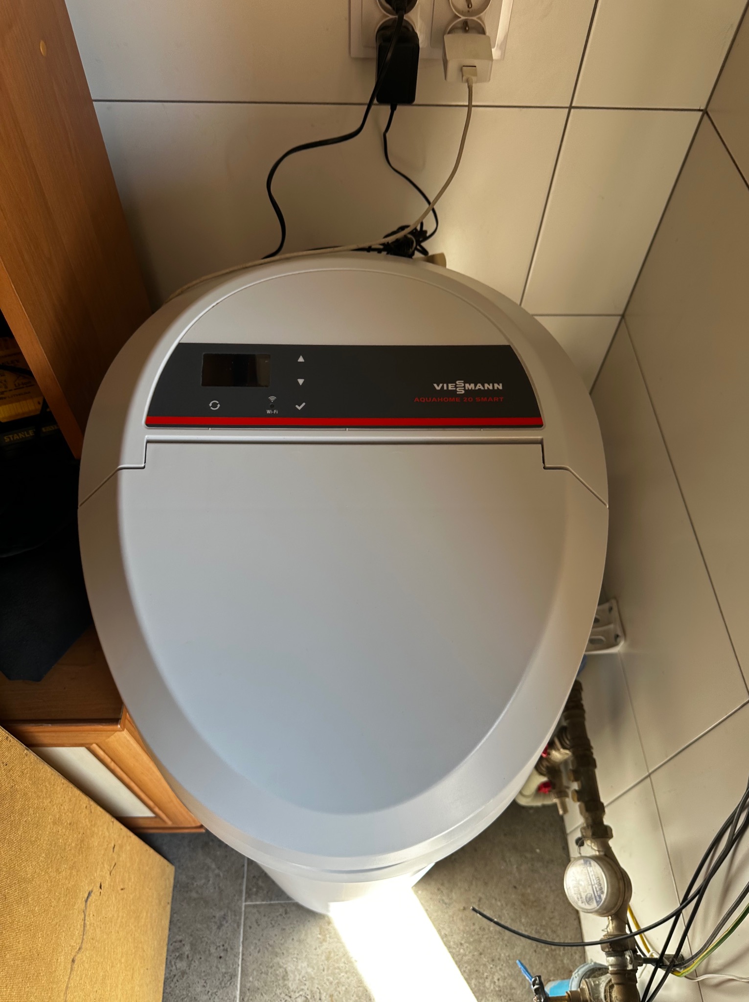 Viessmann Aquahome 20 Smart - zmiękczacz wody. Widoczne przyłącza i przewody elektryczne. Biała obudowa urządzenia na tle płytek.