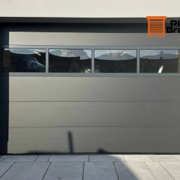 PixBram Dawid Kasprzyk - Szara segmentowa brama garażowa z czterema prostokątnymi oknami, przez które widać drabinę i częściowo samochód. Widoczny fragment elewacji budynku i kostki brukowej.