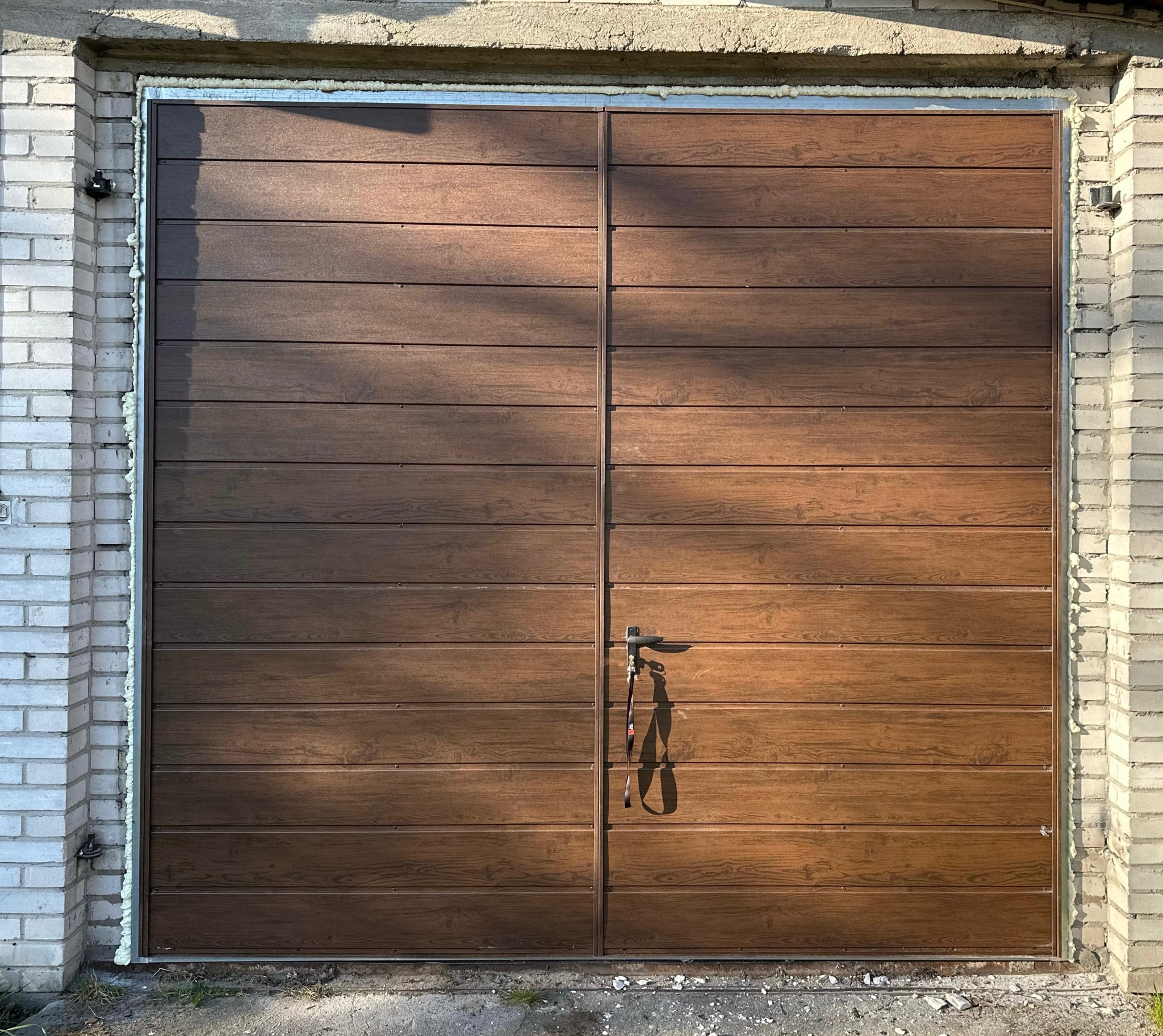 Nowa, brązowa brama garażowa segmentowa z poziomymi panelami imitującymi drewno, zamontowana w murowanym garażu. Widoczne uszczelnienia pianką montażową.
