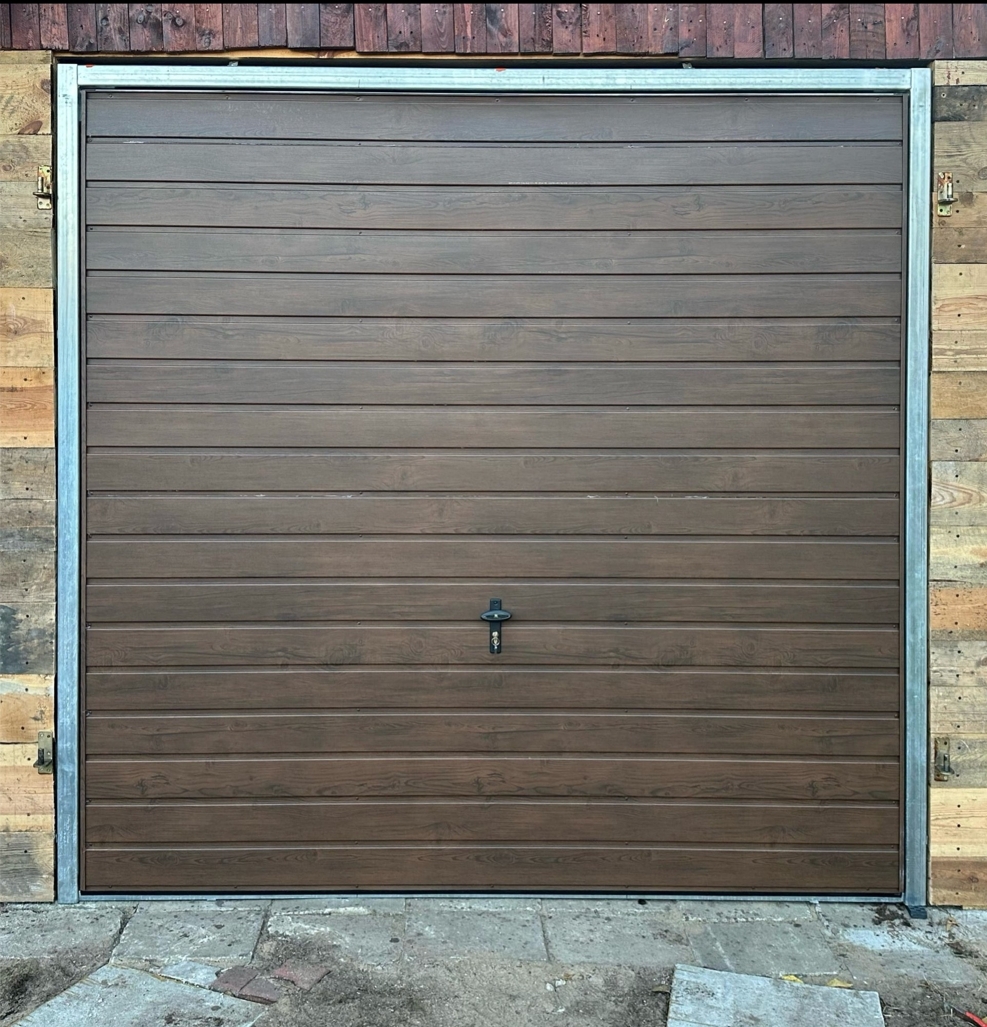 Zamknięta, brązowa brama garażowa z poziomym wzorem drewna, osadzona w metalowej ramie, na tle ściany z desek o zróżnicowanej kolorystyce i fakturze.