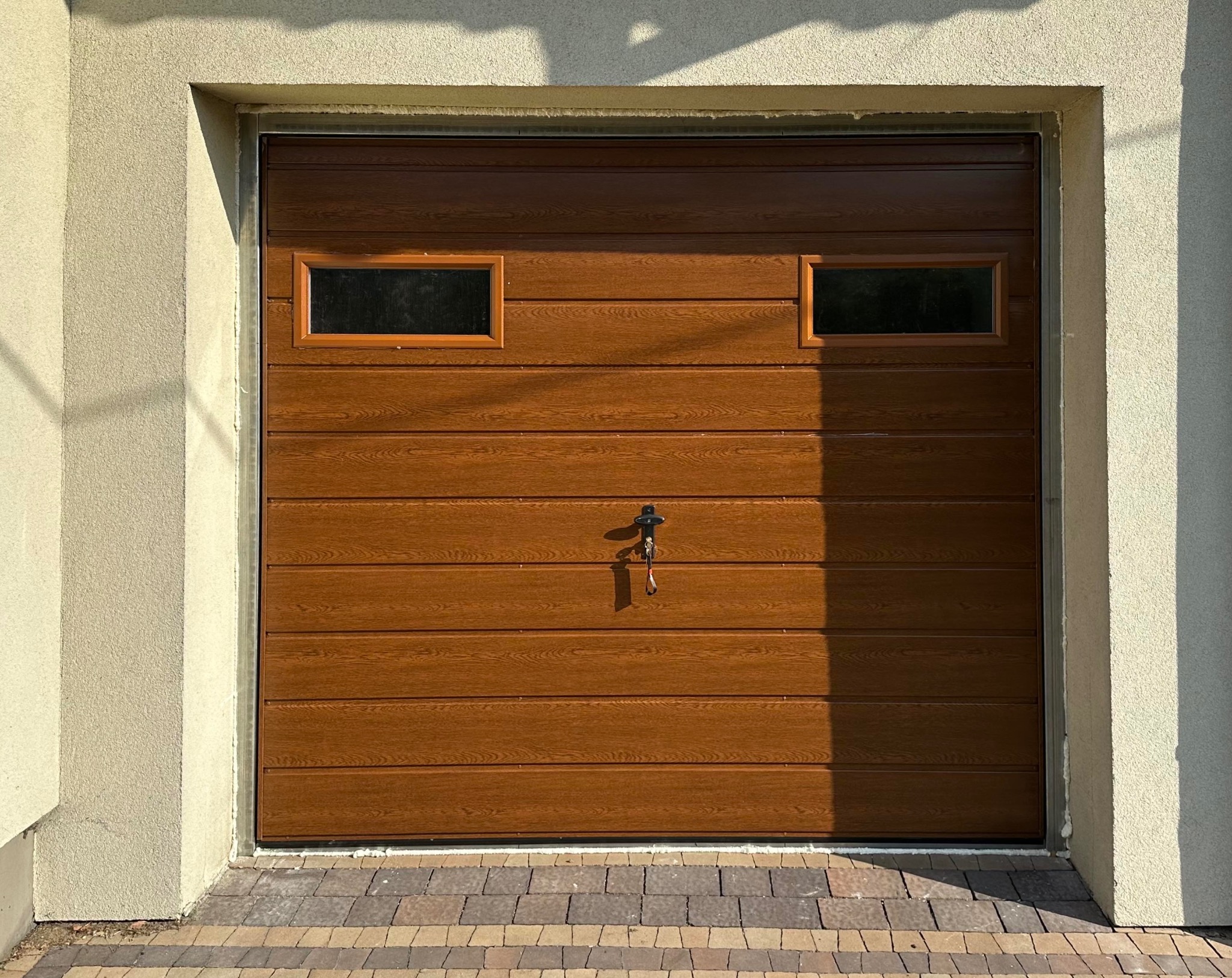 Panelowa brama garażowa w odcieniach brązu z dwoma prostokątnymi okienkami, zamontowana w elewacji budynku. Widoczny zamek z kluczem i cień.