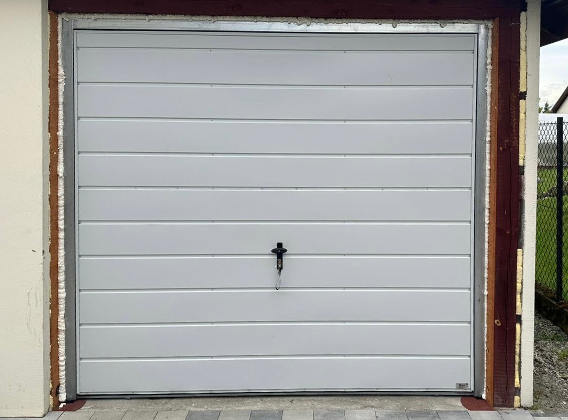 Zamknięta, segmentowa brama garażowa w kolorze jasnoszarym, z widoczną uszczelką i ramą montażową. Brama z poziomymi przetłoczeniami, zamek na środku.
