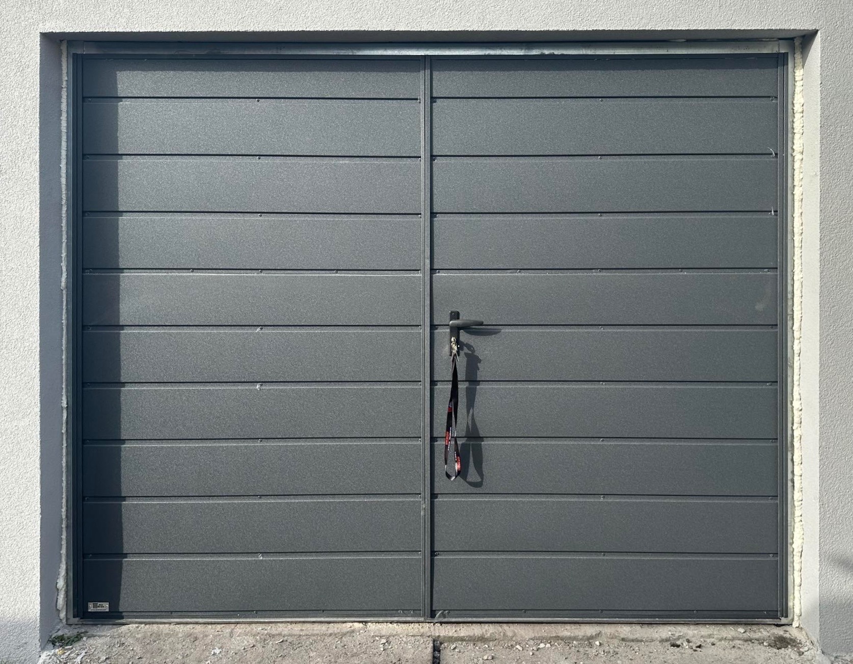 Nowoczesna, dwuskrzydłowa brama garażowa w kolorze grafitowym z poziomymi panelami. Na klamce wiszą klucze. Widoczne uszczelnienie na krawędziach.