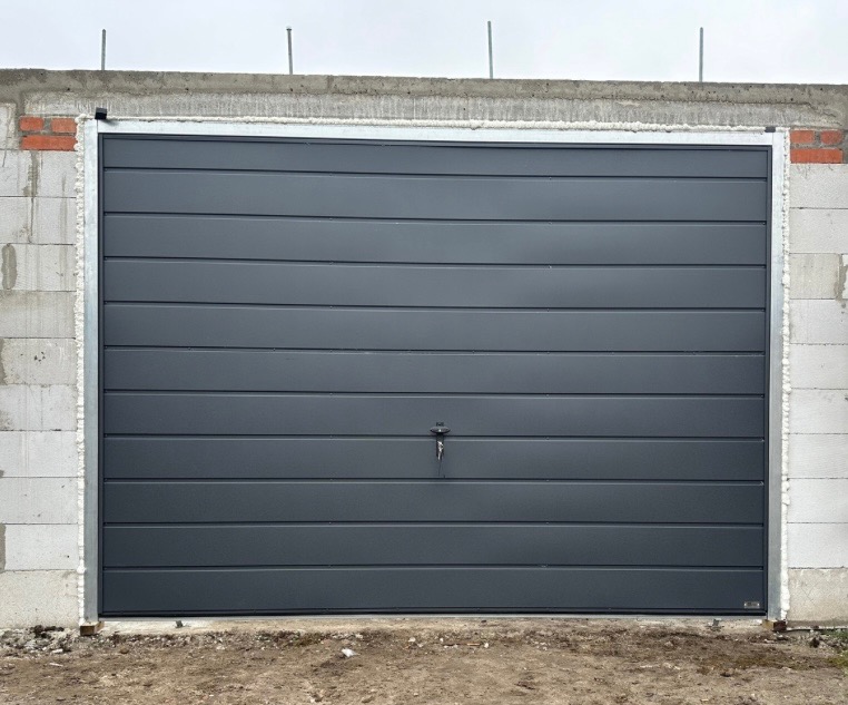 Nowoczesna, segmentowa brama garażowa w kolorze grafitowym, z widocznymi poziomymi przetłoczeniami, zamontowana w surowym otworze z betonu komórkowego.