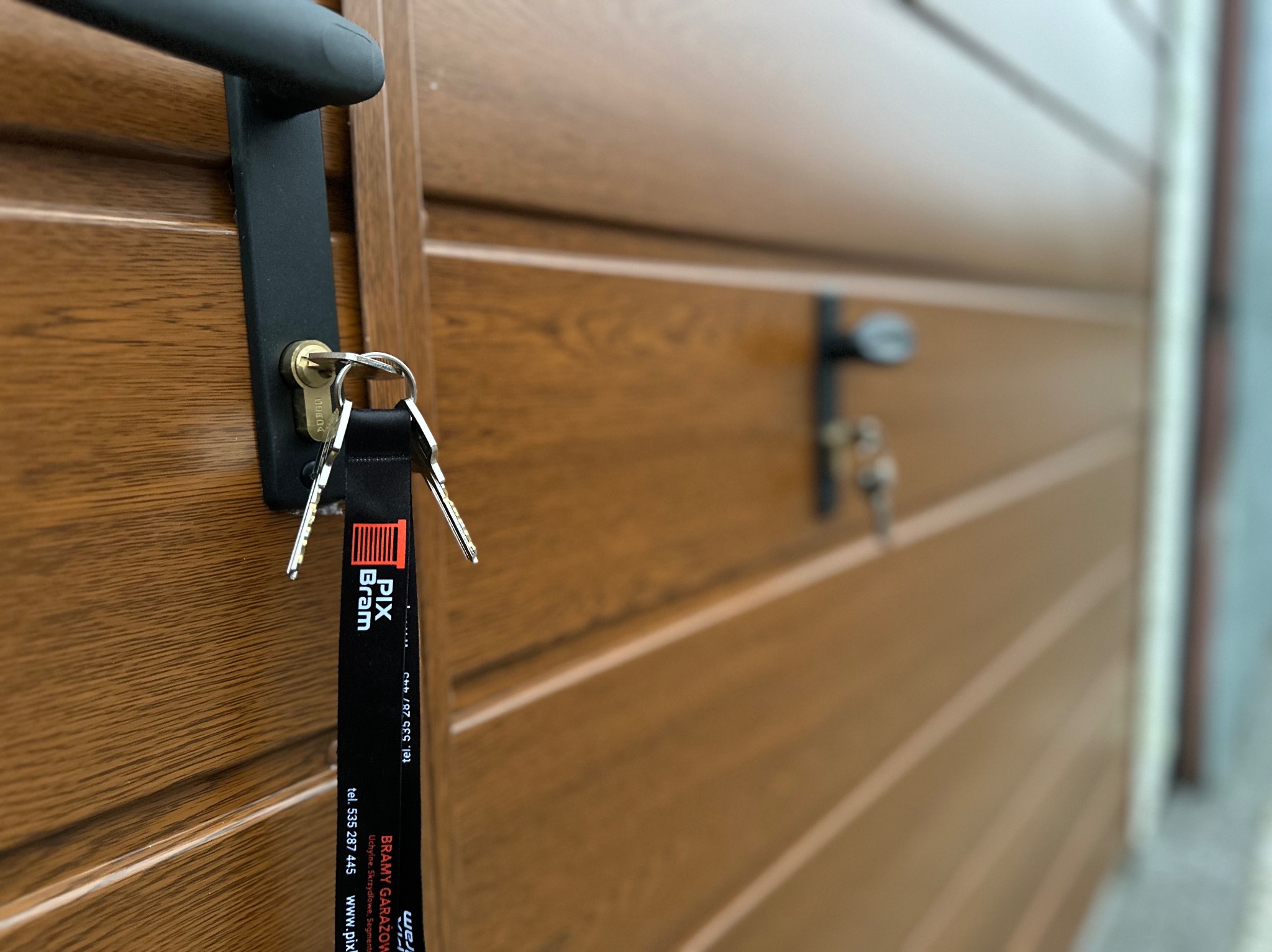 Brązowa brama garażowa segmentowa z czarnym uchwytem i kluczami z zawieszką reklamową firmy PIX Bram.