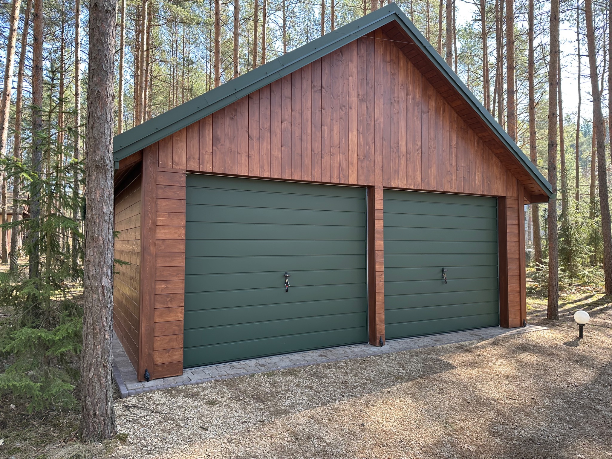 Dwustanowiskowy garaż blaszany z drewnianą elewacją w leśnej scenerii. Zielone bramy segmentowe z kluczami. Oświetlenie zewnętrzne w formie kuli.