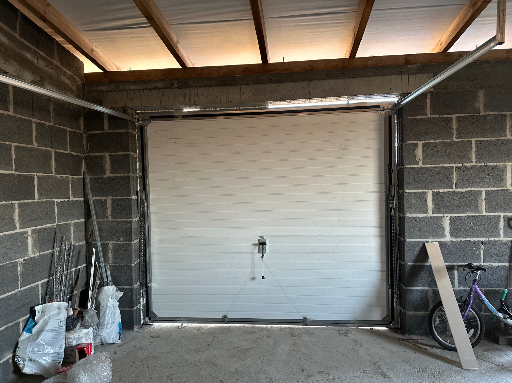 Biała segmentowa brama garażowa w surowym, szarym garażu z betonowych bloczków. Widoczne prowadnice i akcesoria montażowe. W tle drewniana konstrukcja dachu.