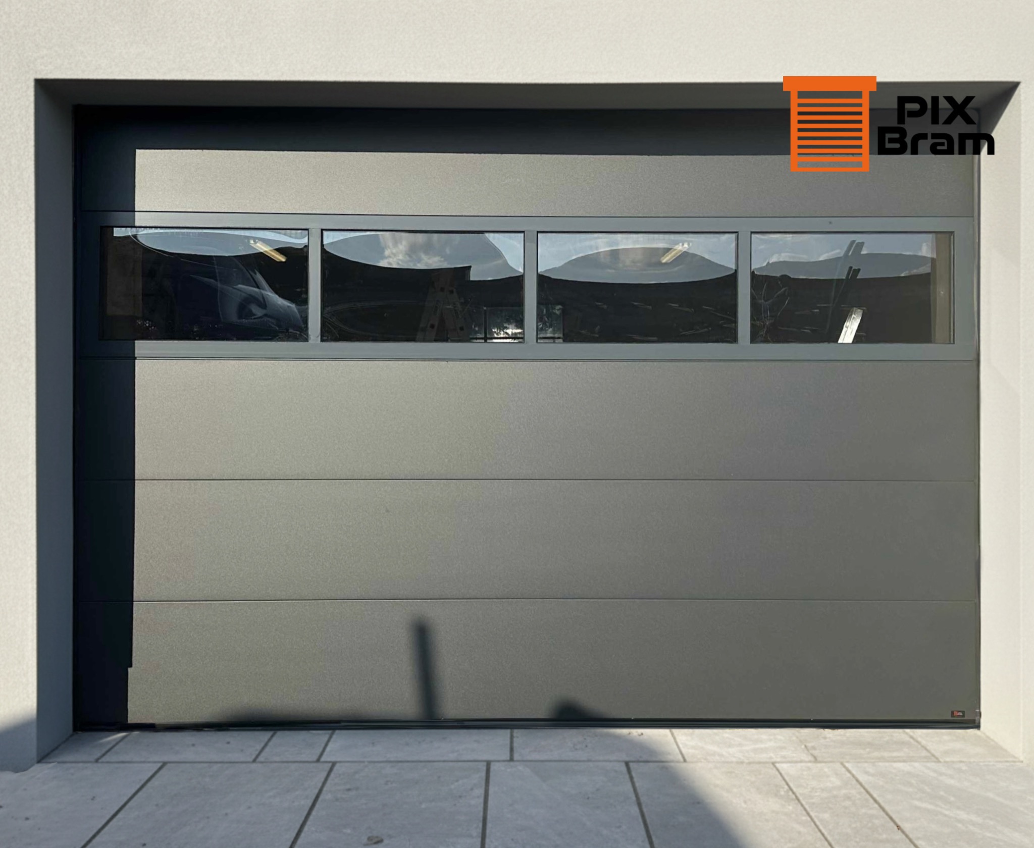 Szara segmentowa brama garażowa z czterema prostokątnymi oknami, przez które widać drabinę i częściowo samochód. Widoczny fragment elewacji budynku i kostki brukowej.