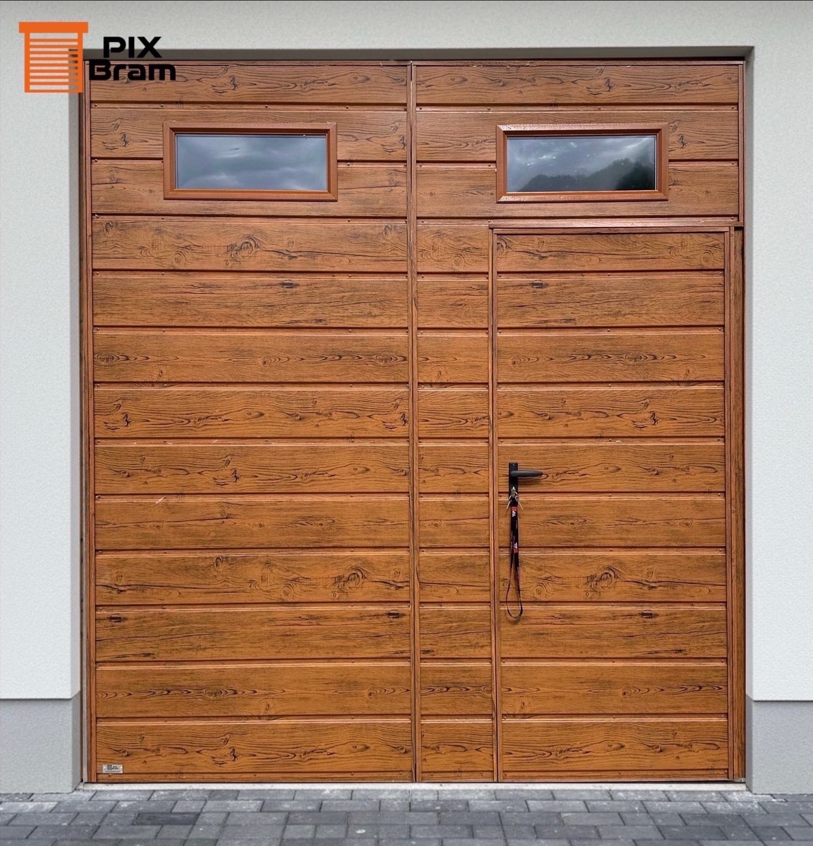 Segmentowa brama garażowa z dwoma oknami i fakturą drewna, czarny uchwyt z breloczkiem, szara kostka brukowa i biała ściana.