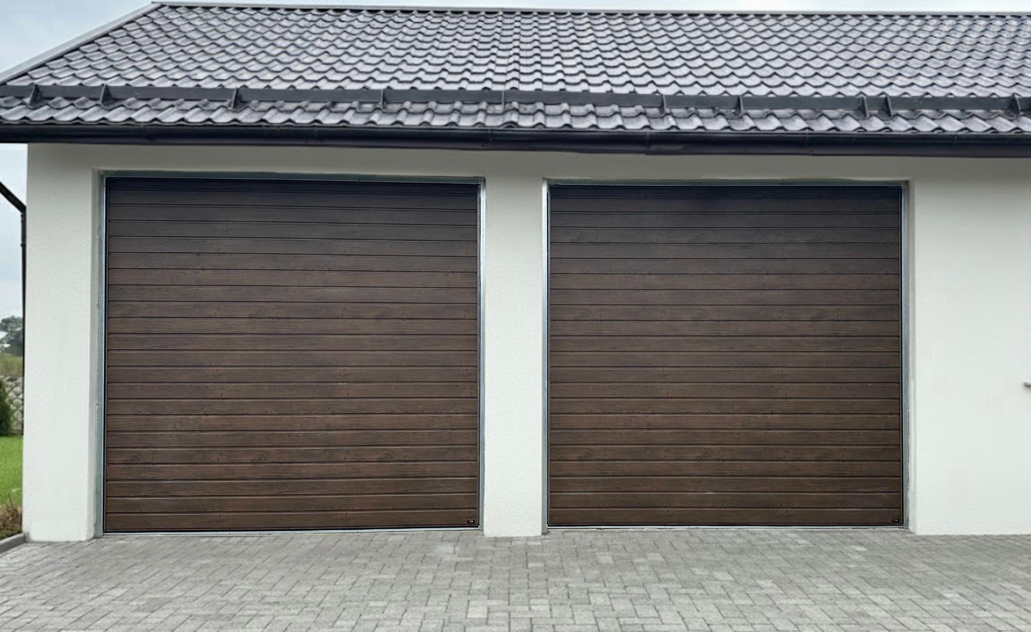 Dwie segmentowe bramy garażowe w kolorze ciemnego drewna, zamontowane w białym budynku z szarą dachówką. Widok z zewnątrz, na pierwszym planie kostka brukowa.
