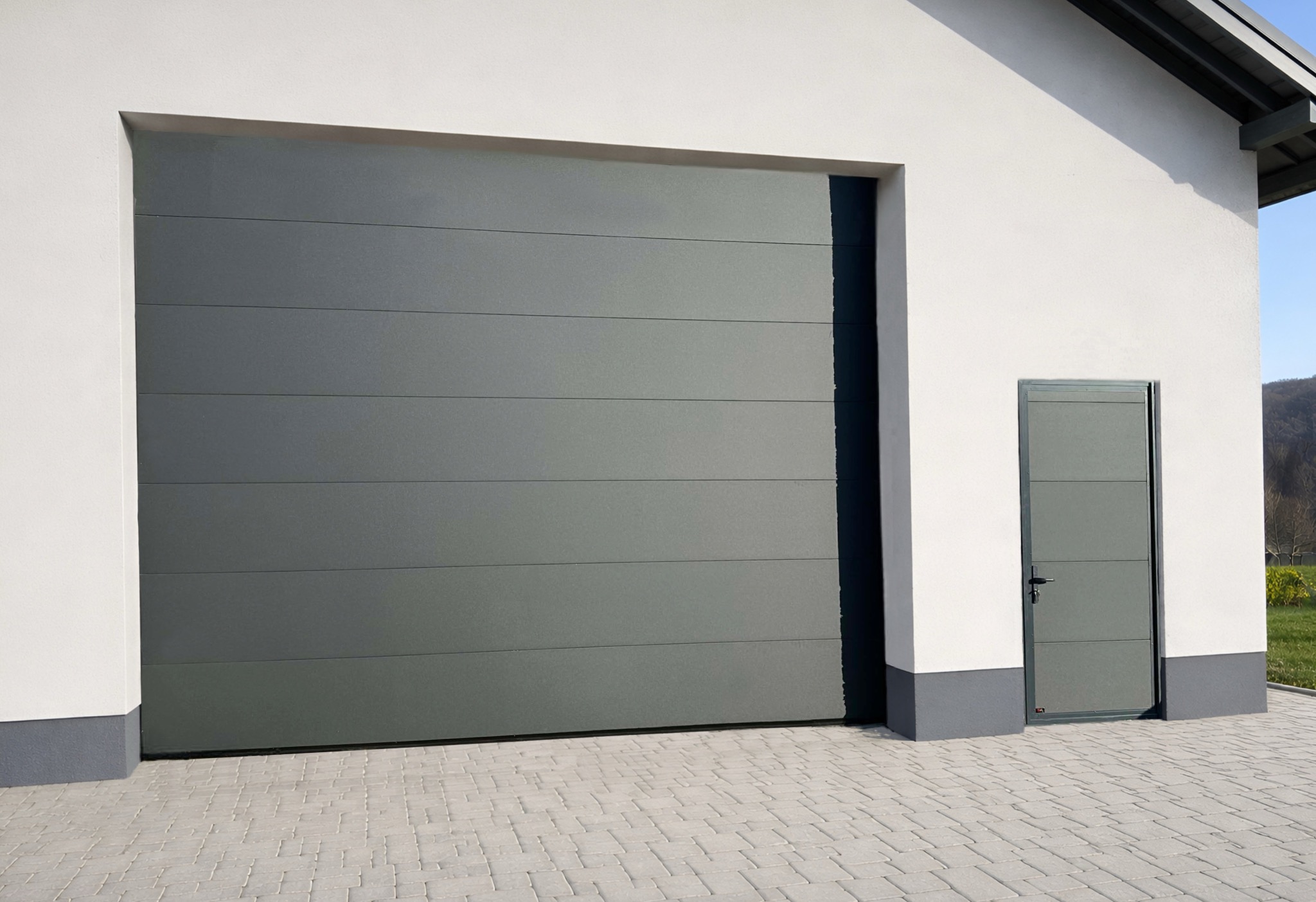 Szara brama garażowa segmentowa z drzwiami bocznymi w nowoczesnym budynku z białymi ścianami i szarą podmurówką, kostka brukowa na podjeździe.