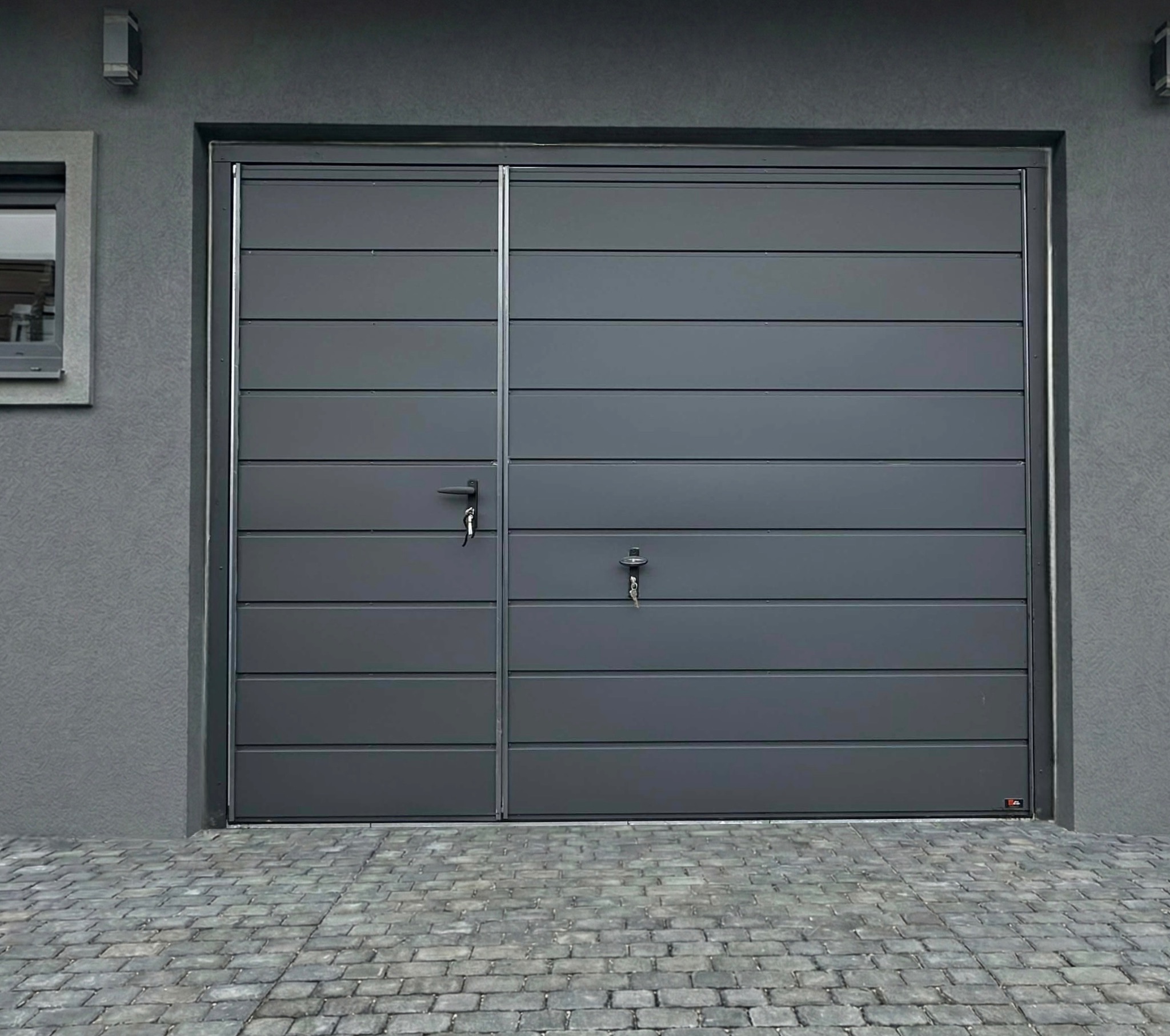 Szara segmentowa brama garażowa z bocznym wejściem i kluczami w zamkach, na tle szarej elewacji i kostki brukowej. Minimalistyczny design.