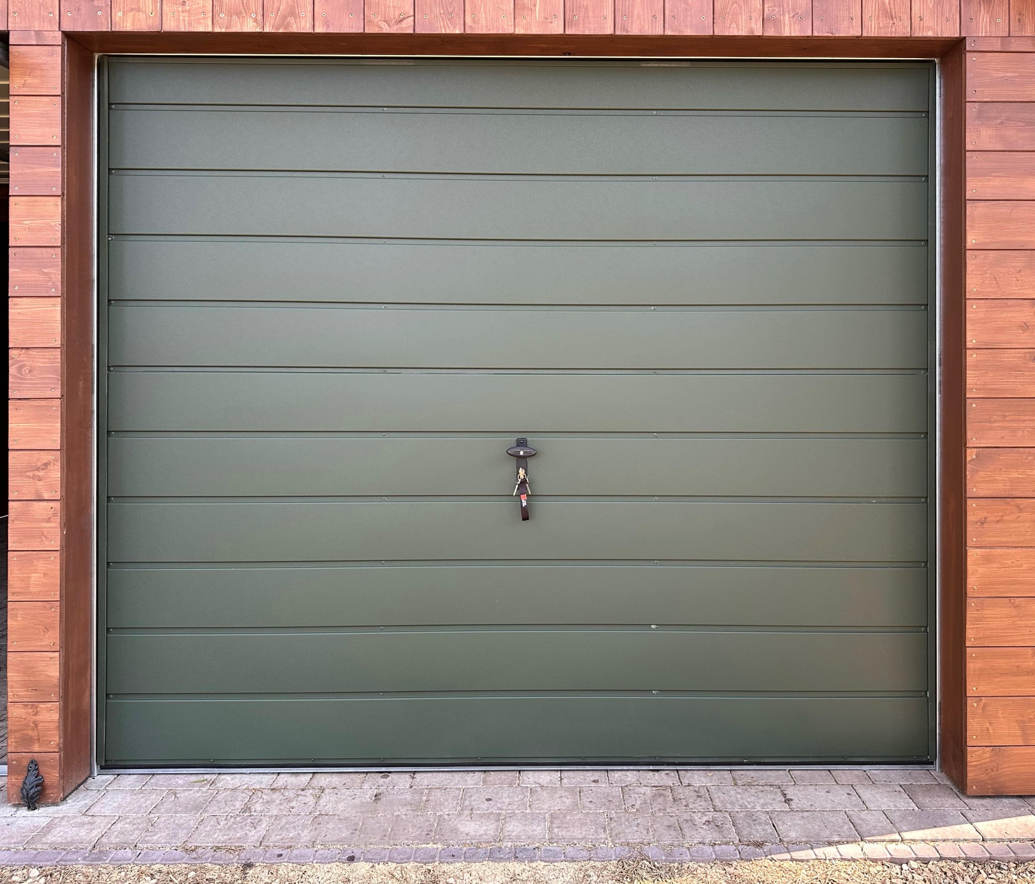 Zamknięta, segmentowa brama garażowa w odcieniach zieleni, z poziomymi przetłoczeniami, zamontowana w drewnianej obudowie. Na bramie wiszą klucze.