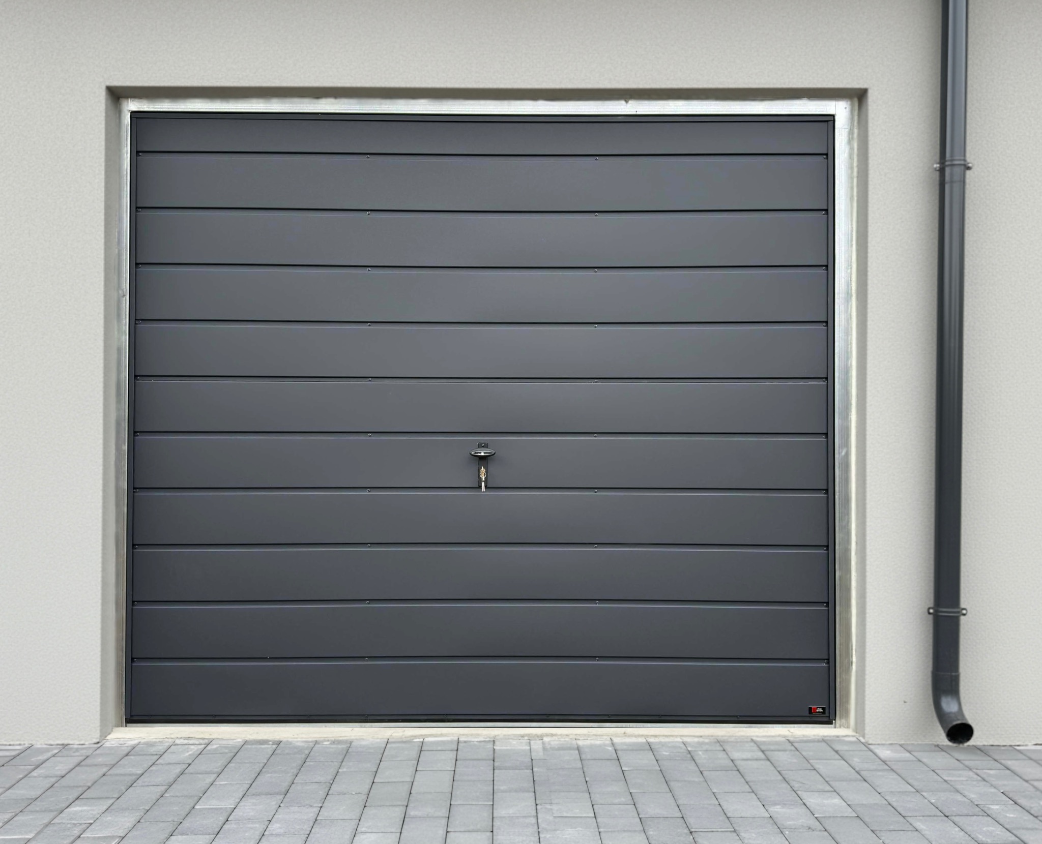 Szara, segmentowa brama garażowa z poziomymi panelami, zamknięta. Widoczny zamek z kluczem, fragment elewacji i rynna. Kostka brukowa przed bramą.