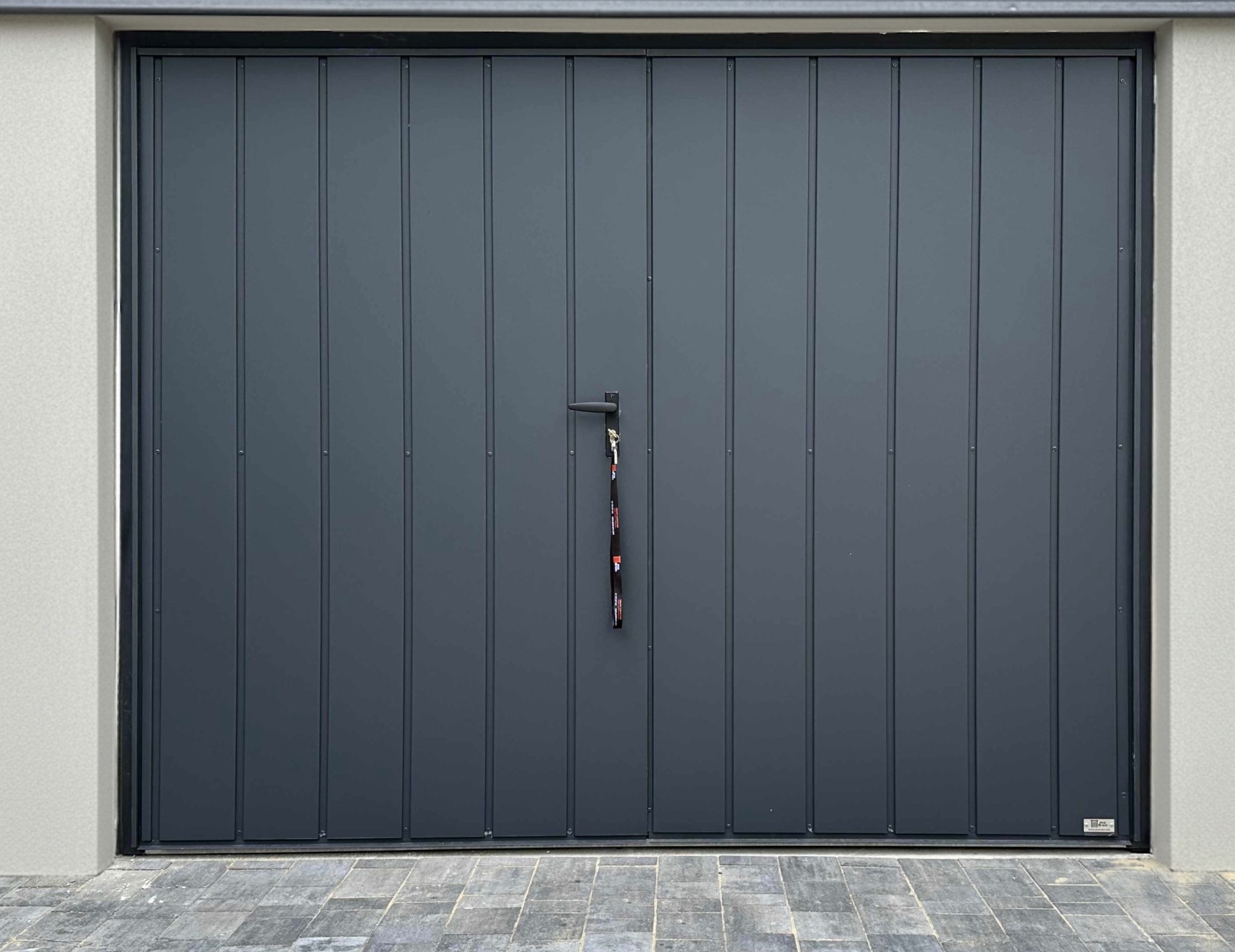 Grafitowa brama garażowa segmentowa z pionowymi przetłoczeniami, z czarną klamką i brelokiem, zamontowana w jasnym murze. Kostka brukowa przed bramą.