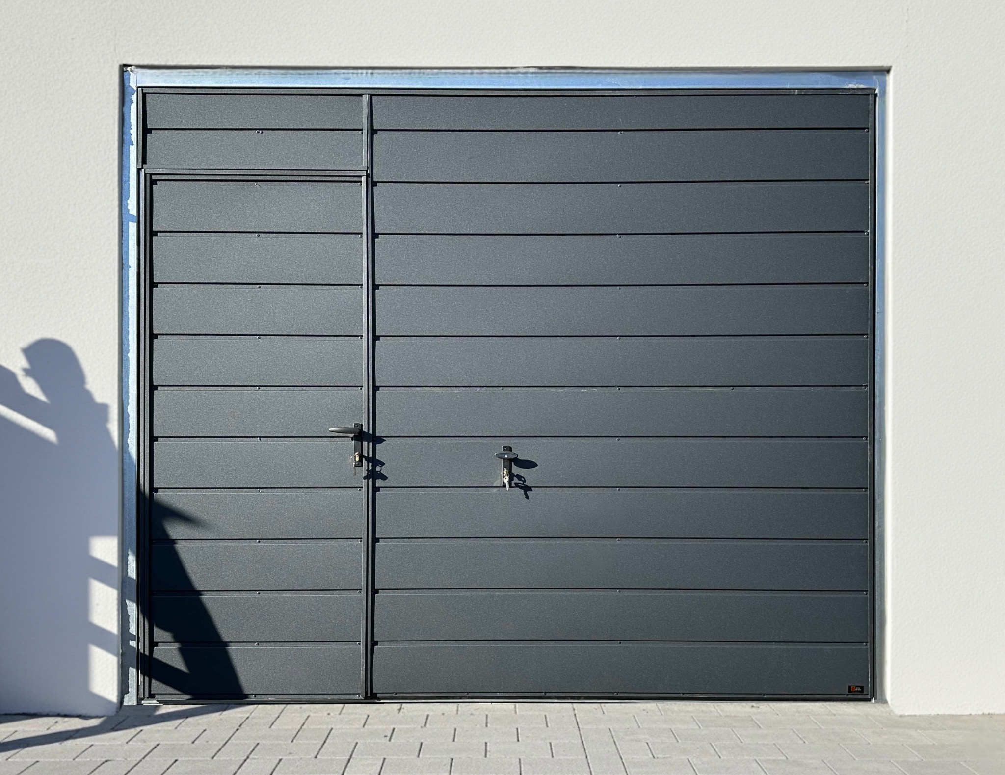 Antracytowa brama garażowa segmentowa z bocznym wejściem, zamykana na dwa zamki, na tle białej ściany i szarej kostki brukowej. Cień osoby po lewej.