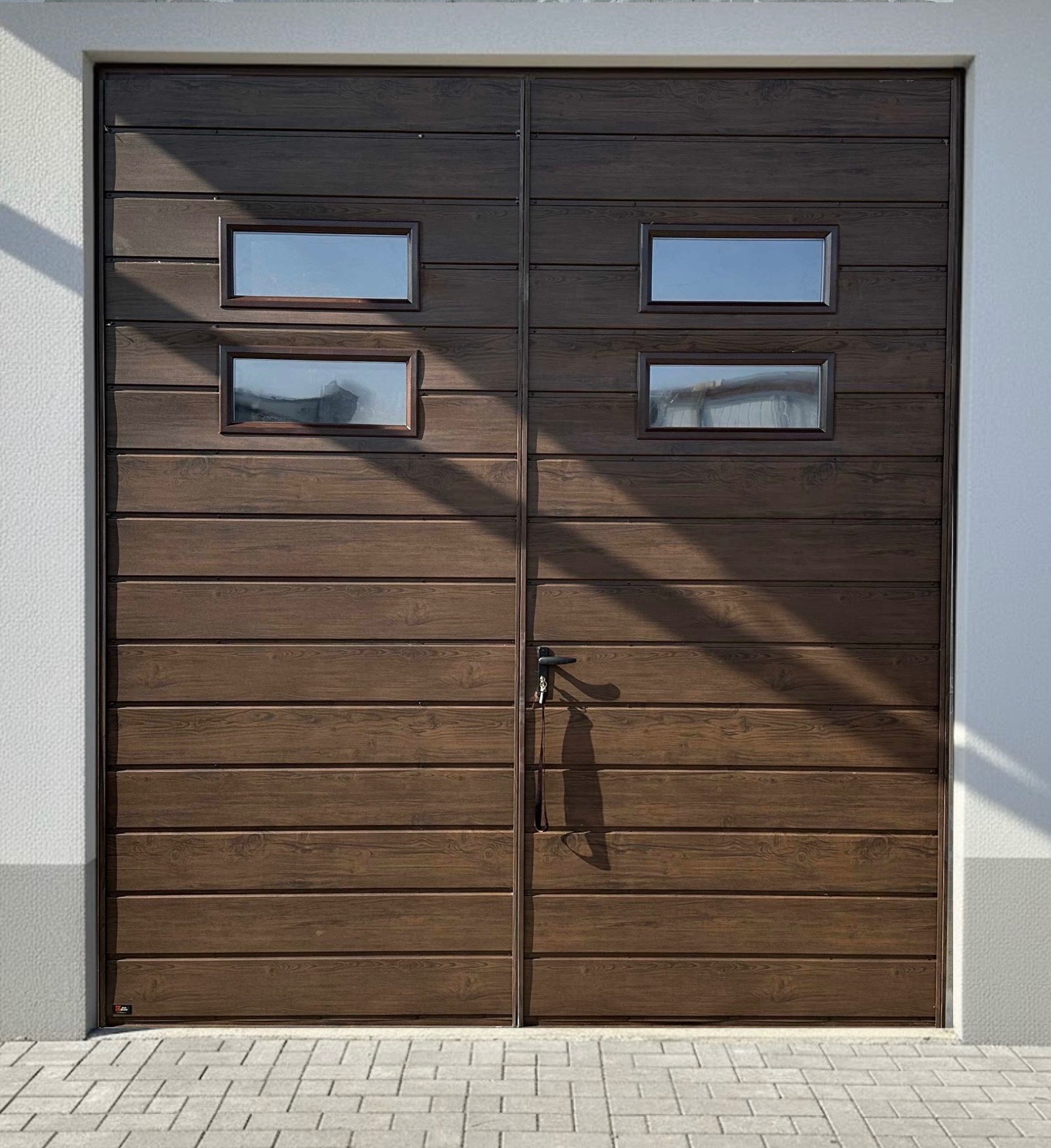 Dwuskrzydłowa brama garażowa z poziomych paneli w odcieniach brązu, z czterema małymi okienkami i czarną klamką, oświetlona promieniami słońca.
