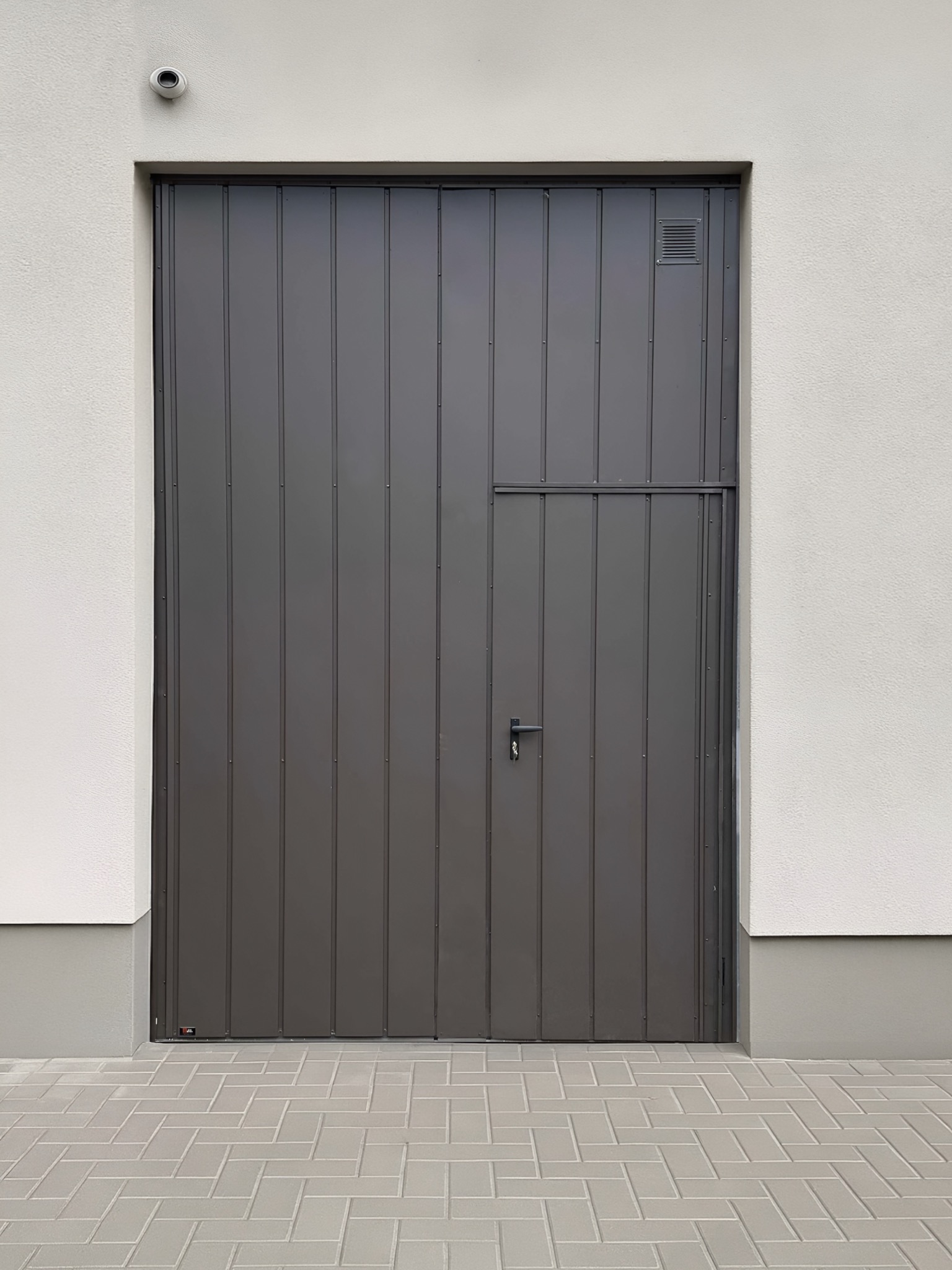Szara brama garażowa segmentowa z małym wejściem, pionowe panele, jasna elewacja budynku, kostka brukowa. Widoczna kamera monitoringu.