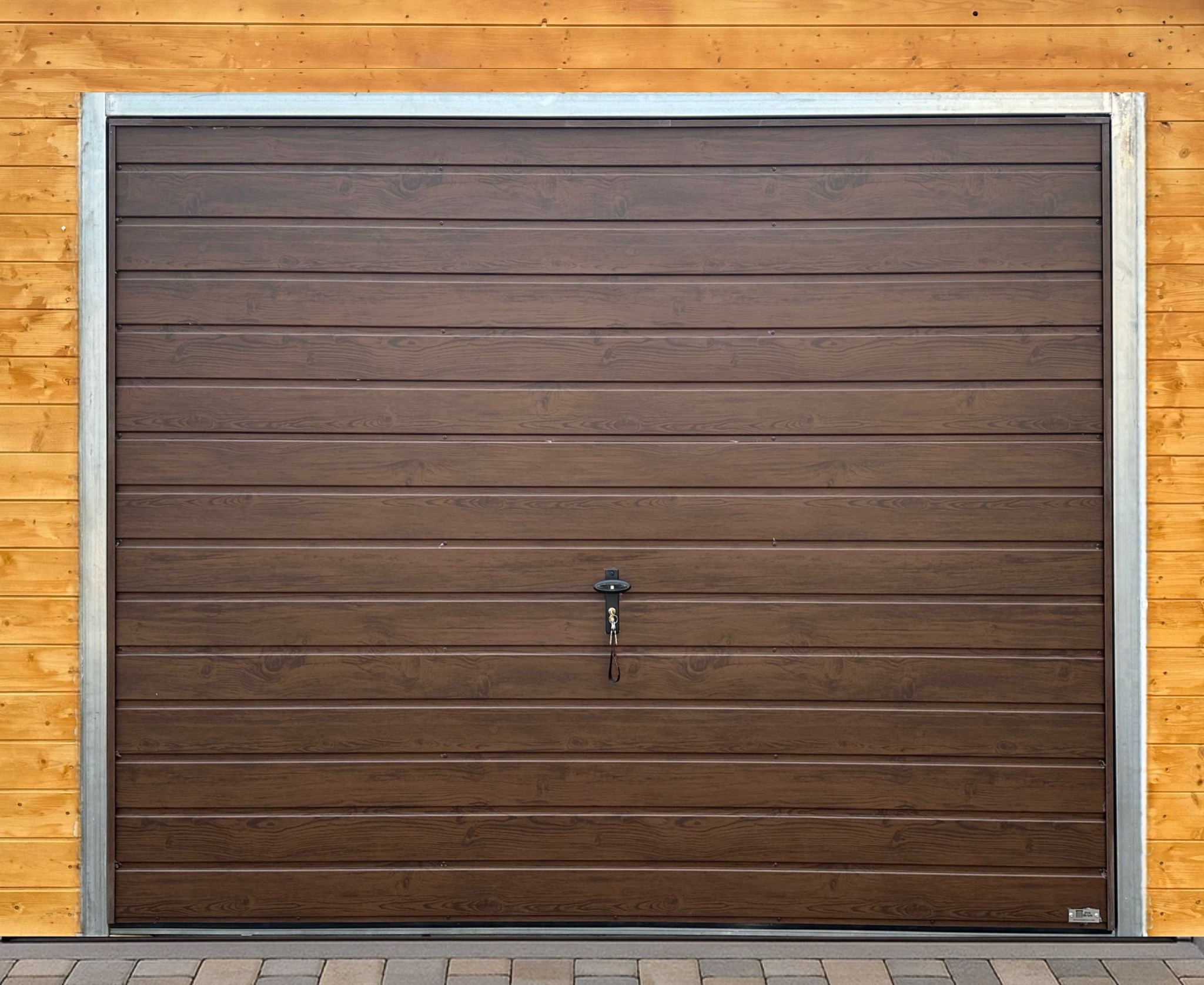 Ciemnobrązowa segmentowa brama garażowa z kluczykiem, zamontowana w drewnianej elewacji. Widoczna metalowa rama i fragment kostki brukowej u dołu.