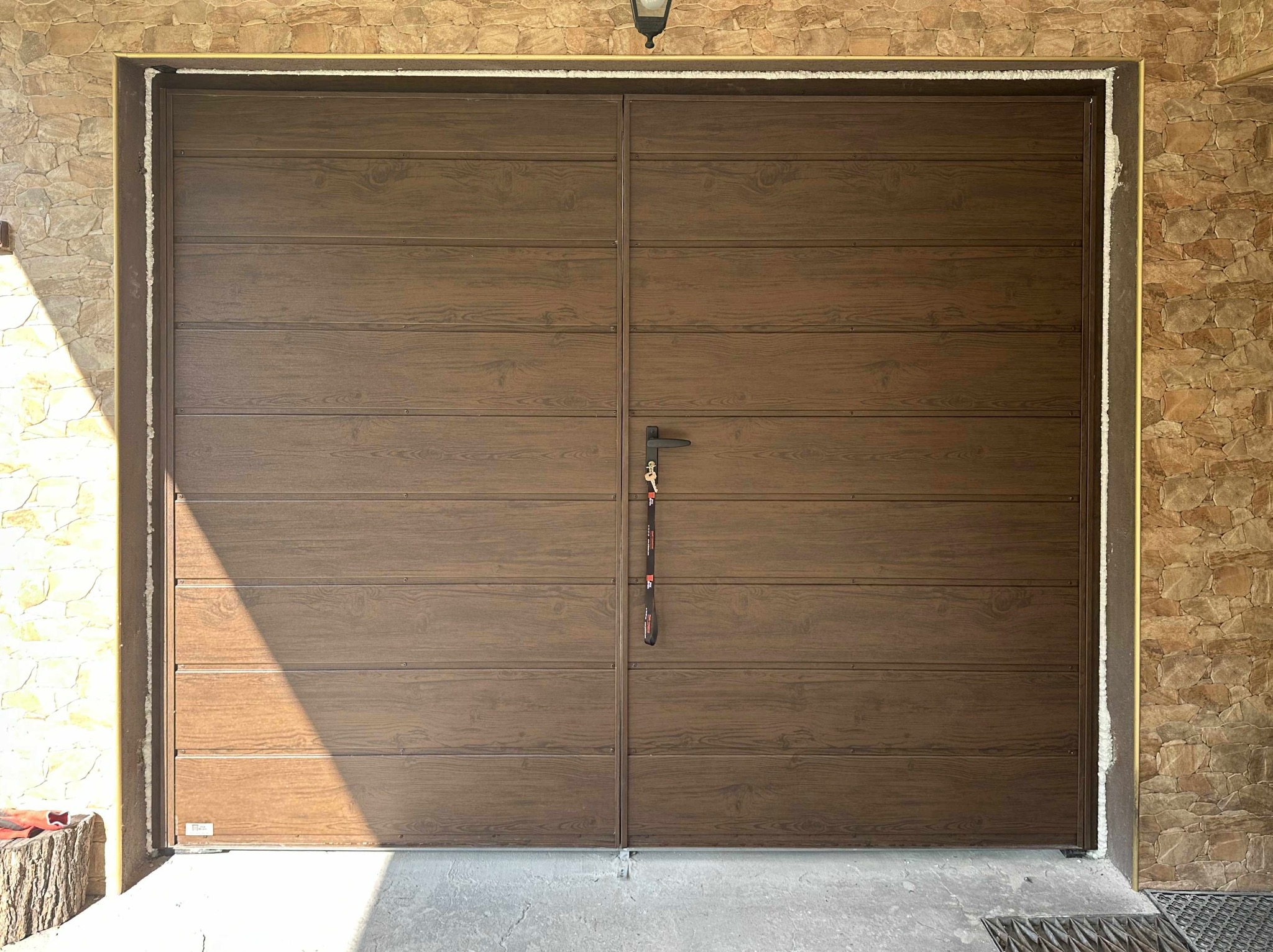 Zamknięta brama garażowa segmentowa w kolorze drewna, z widocznym uszczelnieniem i kluczem w zamku, Wilkowisko. Elewacja z kamienia.