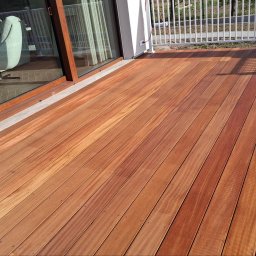 DecorCraft MACIEJ BIŚ - Nowoczesny taras z desek kompozytowych w ciepłym odcieniu brązu, z widocznym fragmentem balustrady i przeszkleniem okiennym. Widoczne pudełka po panelach.