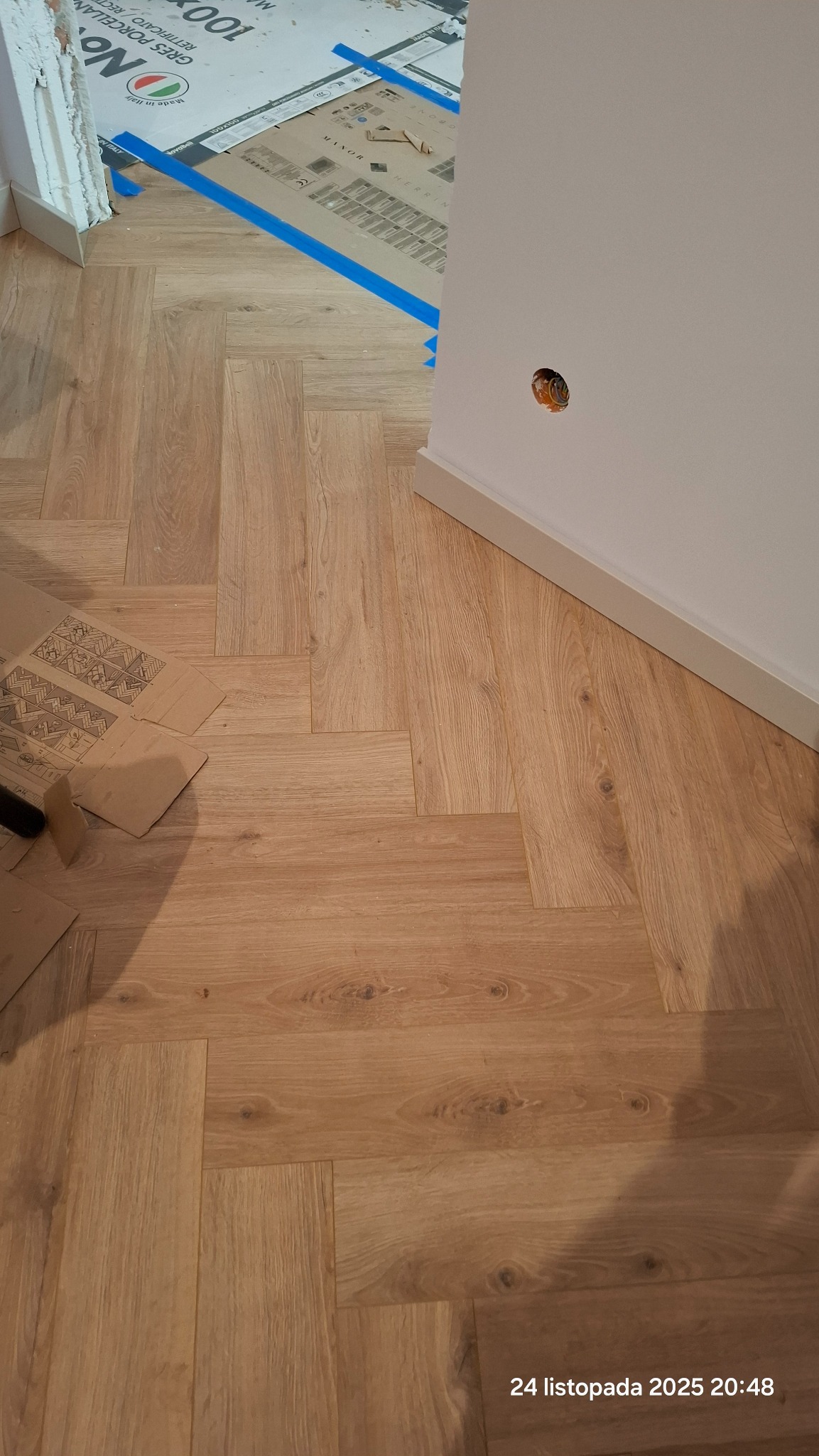 Jasne panele ułożone we wzór jodełki francuskiej, widoczne kartony z oznaczeniami producenta i fragment ściany z gniazdkiem elektrycznym.