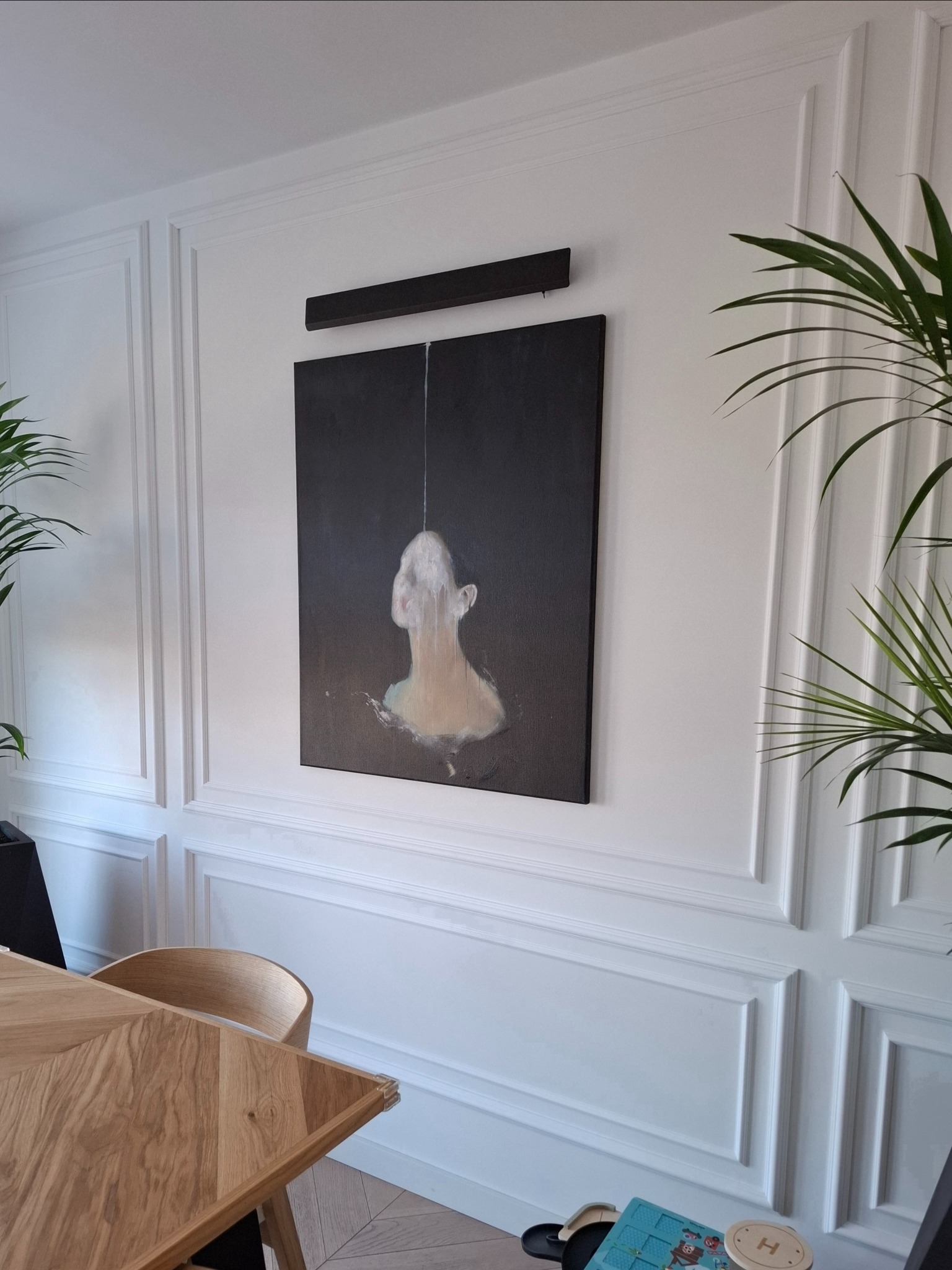 Nowoczesne wnętrze z obrazem na ścianie z dekoracyjną sztukaterią. W tle widoczny stół z drewna i roślina doniczkowa, tworzące spójną aranżację.