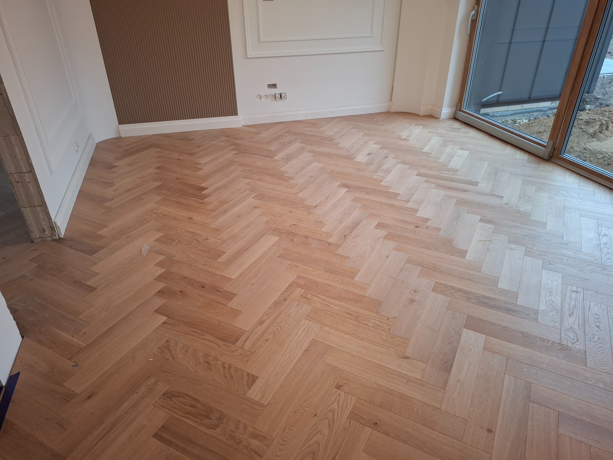 Jasna, dębowa podłoga ułożona w jodełkę francuską w nowym wnętrzu. Widoczne listwy przypodłogowe, ściany w odcieniach bieli i beżu oraz duże okno.