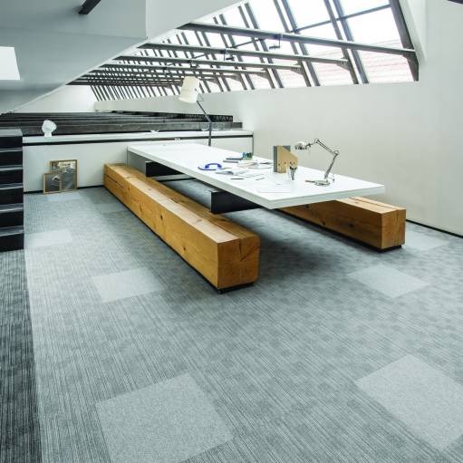 Wnętrze biurowe z szarą wykładziną w kwadraty i drewnianymi ławami pod białym stołem. Duże okna dachowe w tle. Nowoczesny design.