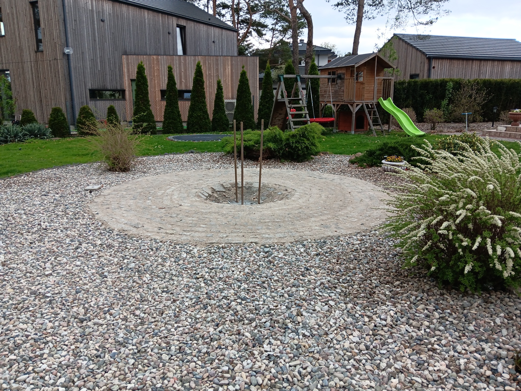 Ogród z wyznaczonym miejscem na ognisko, otoczonym dekoracyjnym kamieniem. W tle widoczne drzewa, trawnik i plac zabaw z domkiem i zjeżdżalnią.