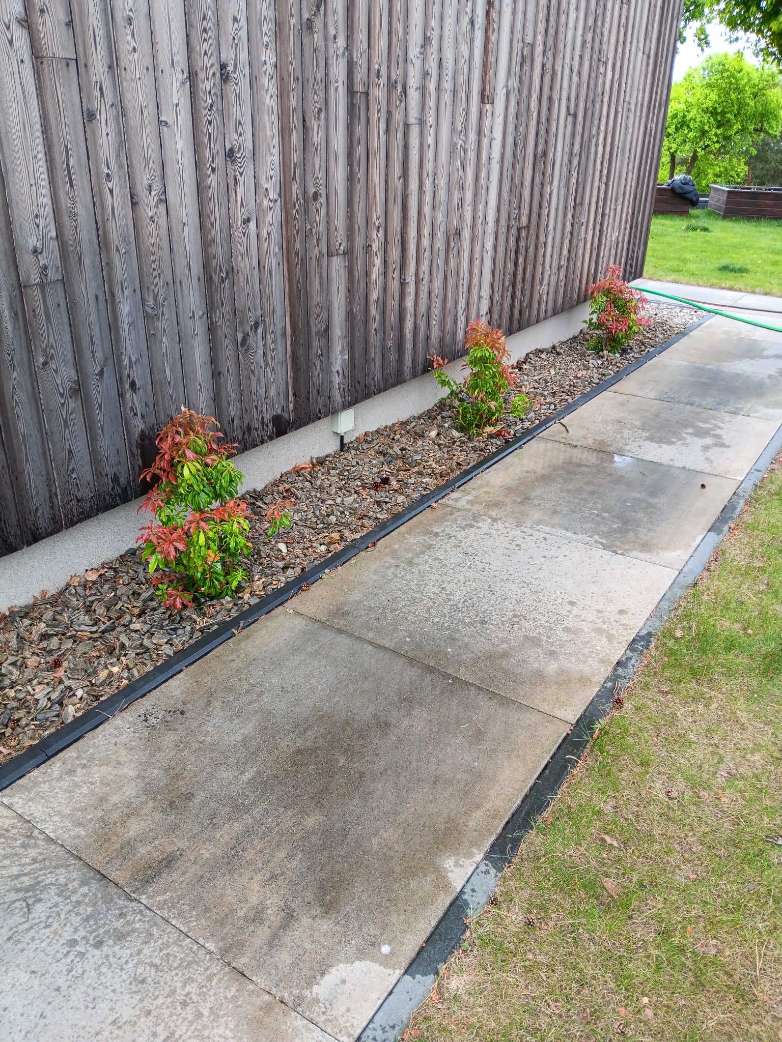 Ścieżka z betonowych płyt obok drewnianej ściany budynku i rabaty z krzewami ozdobnymi, wyłożonej korą. Widoczne detale wykończenia i zieleń.