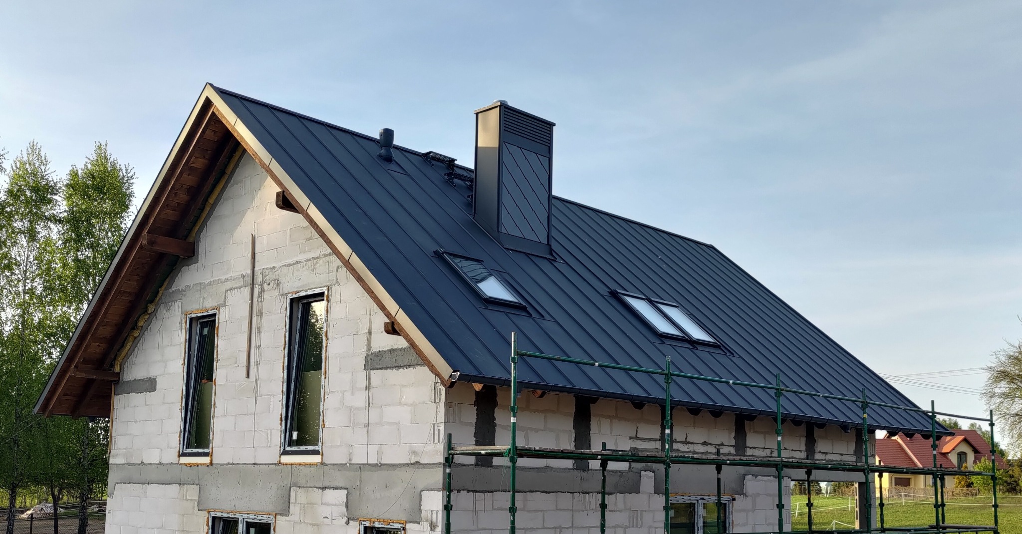 Dom w trakcie budowy z czarnym dachem z blachy na rąbek stojący, kominem i oknami dachowymi. Widoczne rusztowania i surowy stan ścian.