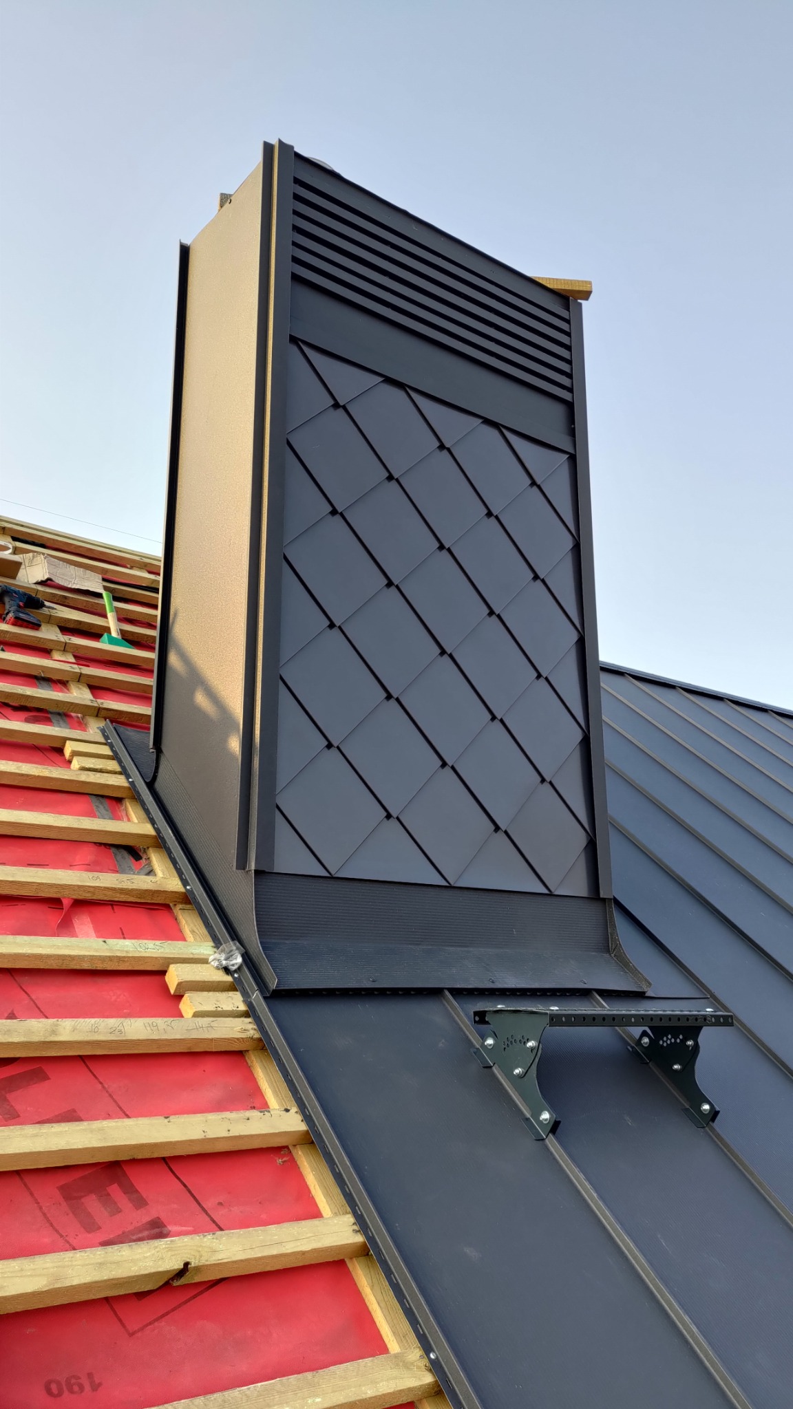 Czarny komin wentylacyjny z romboidalnym wzorem na dachu z blachy na rąbek stojący, widoczne elementy konstrukcji dachu i akcesoria montażowe.