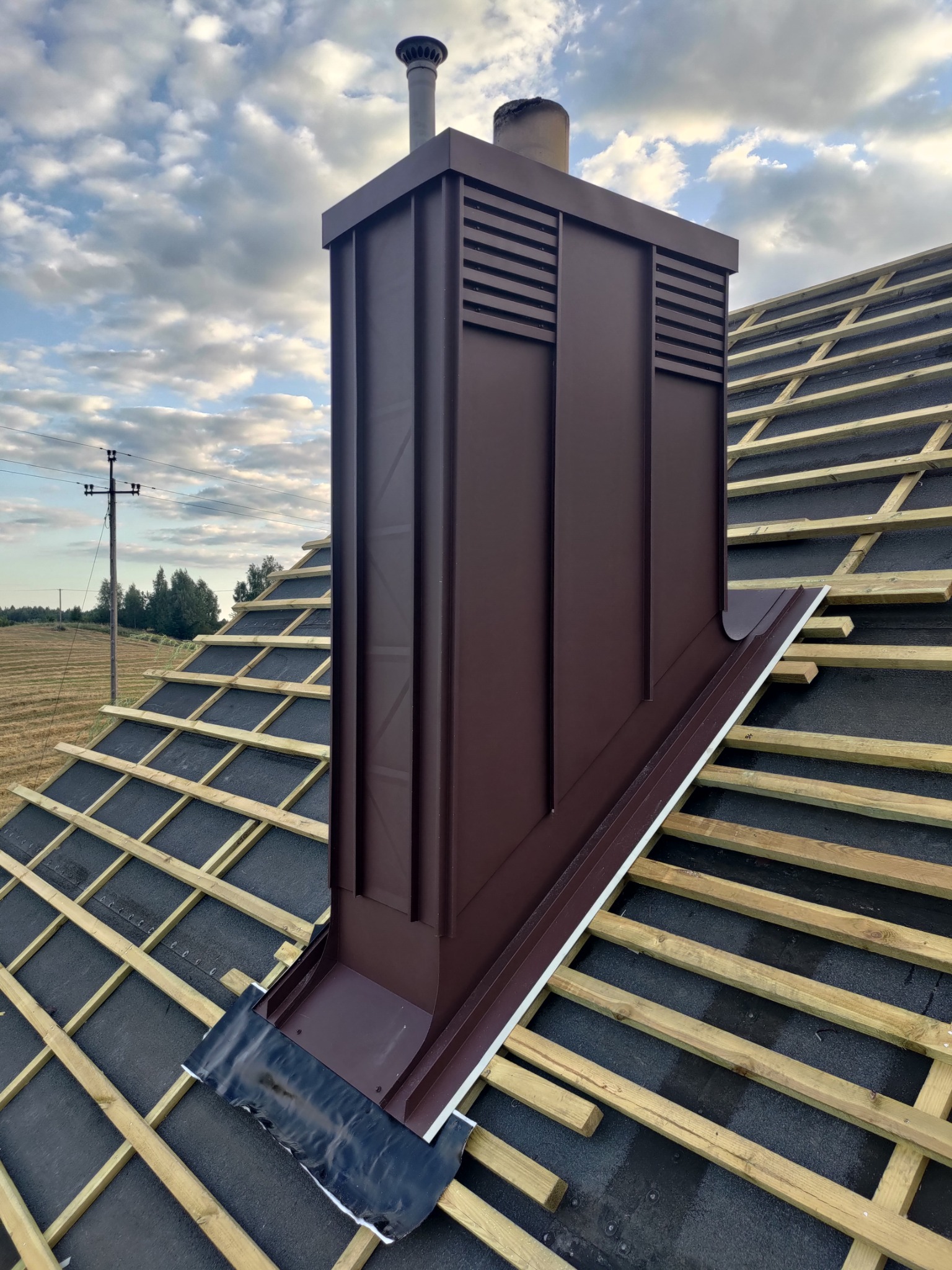 Nowy, brązowy komin wentylacyjny z blachy na dachu z łatami i papą. Widok z dołu, na tle błękitnego nieba z chmurami i pola w oddali. Detal wykonania.