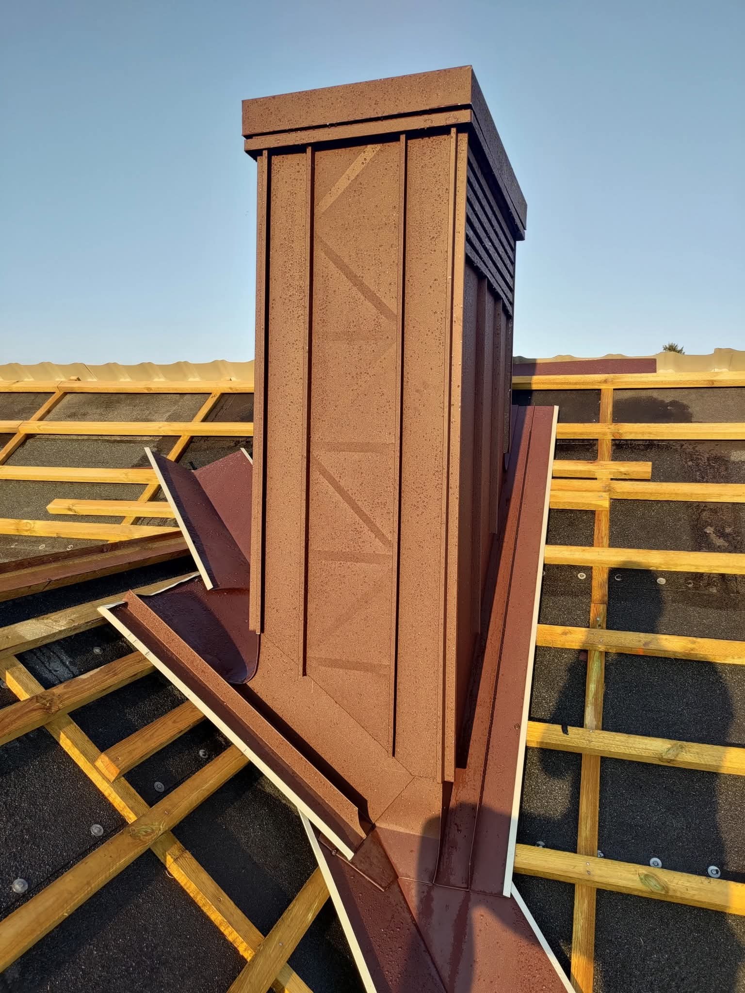 Brązowy komin wentylacyjny na dachu z drewnianą konstrukcją i papą. Widoczne detale obróbek blacharskich i elementy wentylacyjne. Ujęcie z góry, w słoneczny dzień.