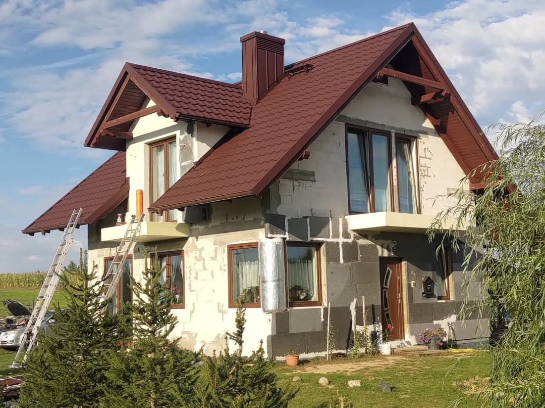 Dom w trakcie ocieplania z brązowym dachem z blachodachówki. Widoczne rusztowania, częściowo ocieplona elewacja i fragment krajobrazu w tle.