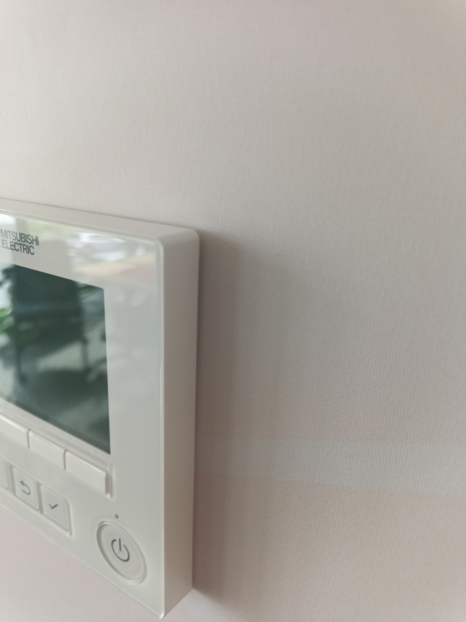 Biały panel sterowania klimatyzacji Mitsubishi Electric zamontowany na ścianie pokrytej jasną tapetą o subtelnej strukturze.