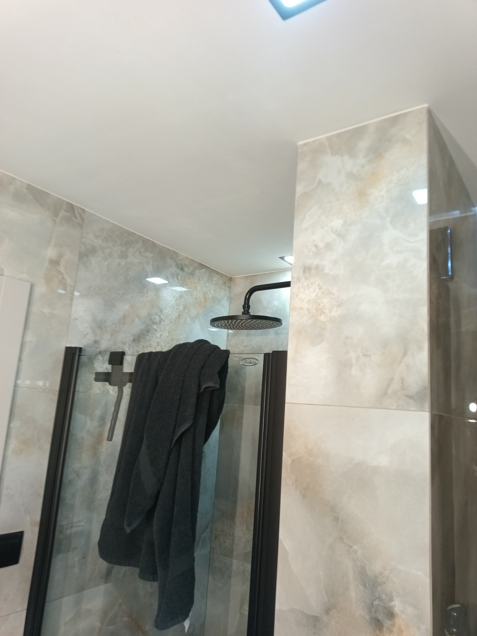 Wykończona kabina prysznicowa z gresem szkliwionym, czarną armaturą i szklaną ścianką działową. Widoczny ręcznik i oświetlenie sufitowe.