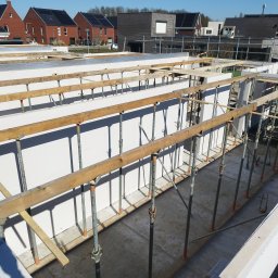 WIKO BOUW Grzegorz Serafin - Wznoszenie ścian z białych bloczków, wspartych stemplami budowlanymi na fundamencie. W tle zabudowa jednorodzinna, słoneczny dzień.