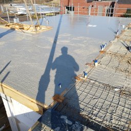 WIKO BOUW Grzegorz Serafin - Wylany beton na stropie z widocznym zbrojeniem i shadow person. Prace budowlane w toku, drewniane szalunki i elementy konstrukcyjne. Dom w tle.