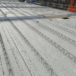 WIKO BOUW Grzegorz Serafin - Zbrojenie stropu Teriva z widocznymi belkami i pustakami, przygotowane do zalania betonem. Na drugim planie budynki i żuraw budowlany.